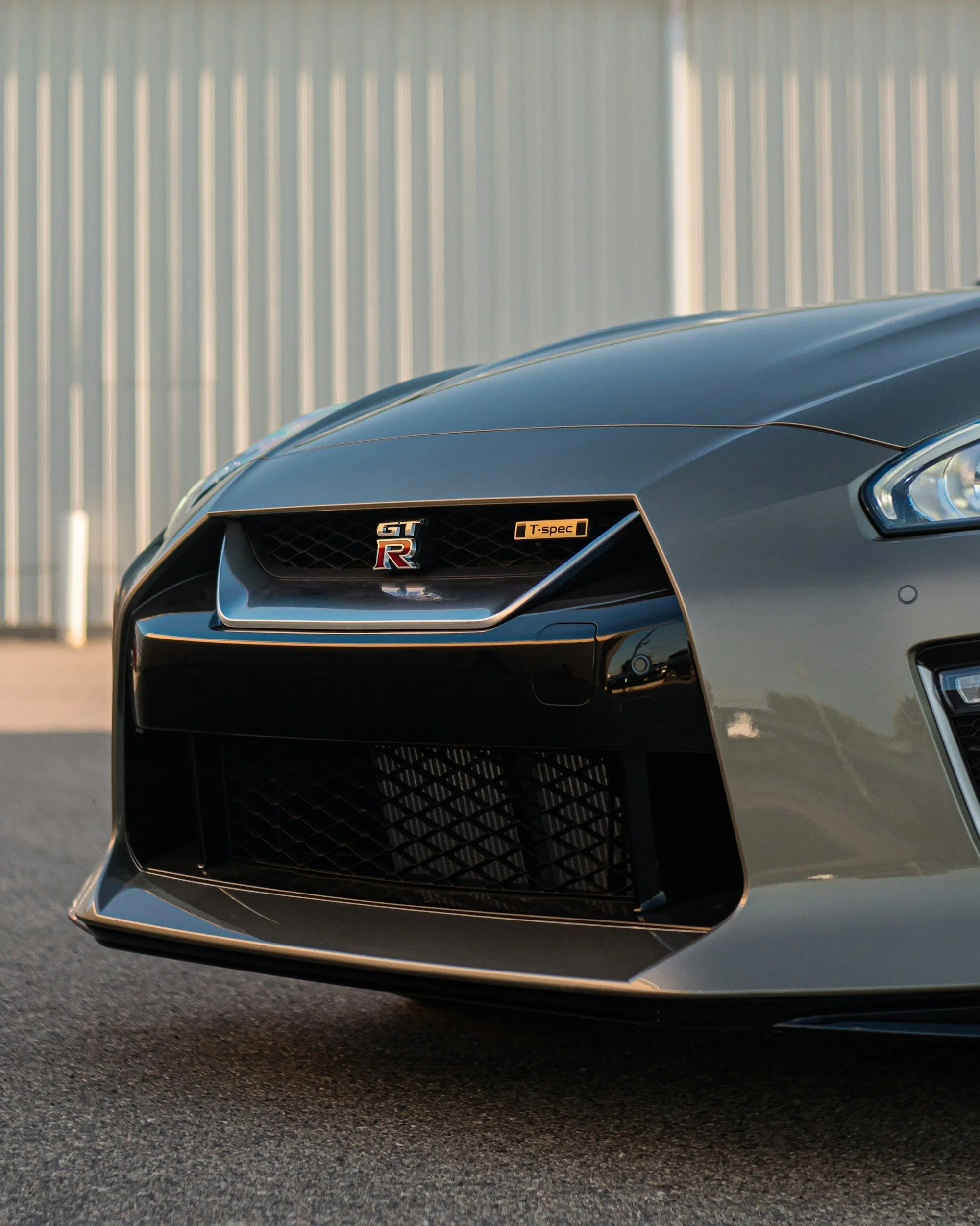 Close-up of the front end of a gray Nissan GT-R sports car, focusing on the grille, badge, and headlight, with a metallic fence in the background.