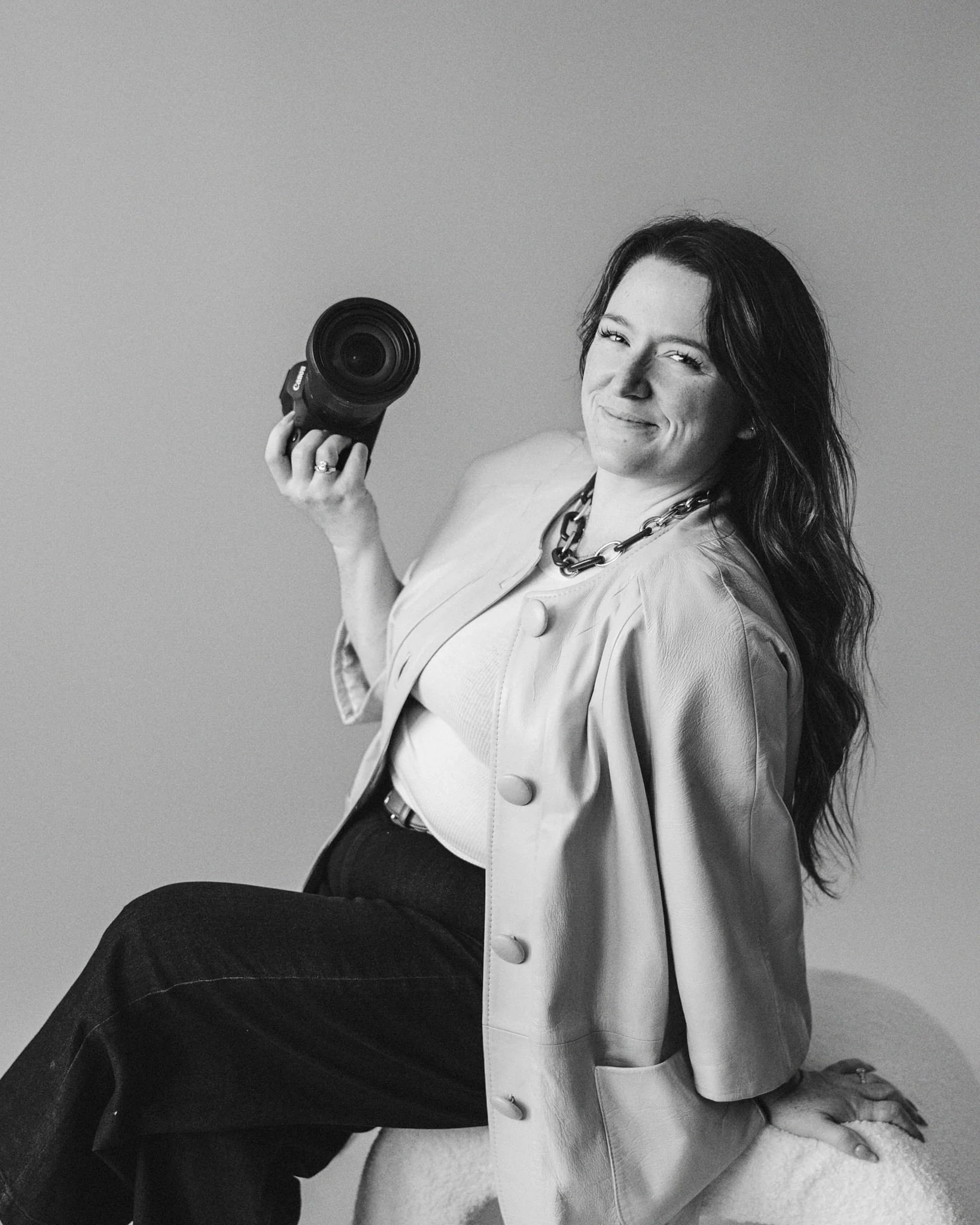 A black and white photo of a woman smiling while holding a camera. She is wearing a light-colored jacket and a chunky necklace, with long hair. The woman appears to be sitting on the floor with her knees bent and one arm resting on her knee.
