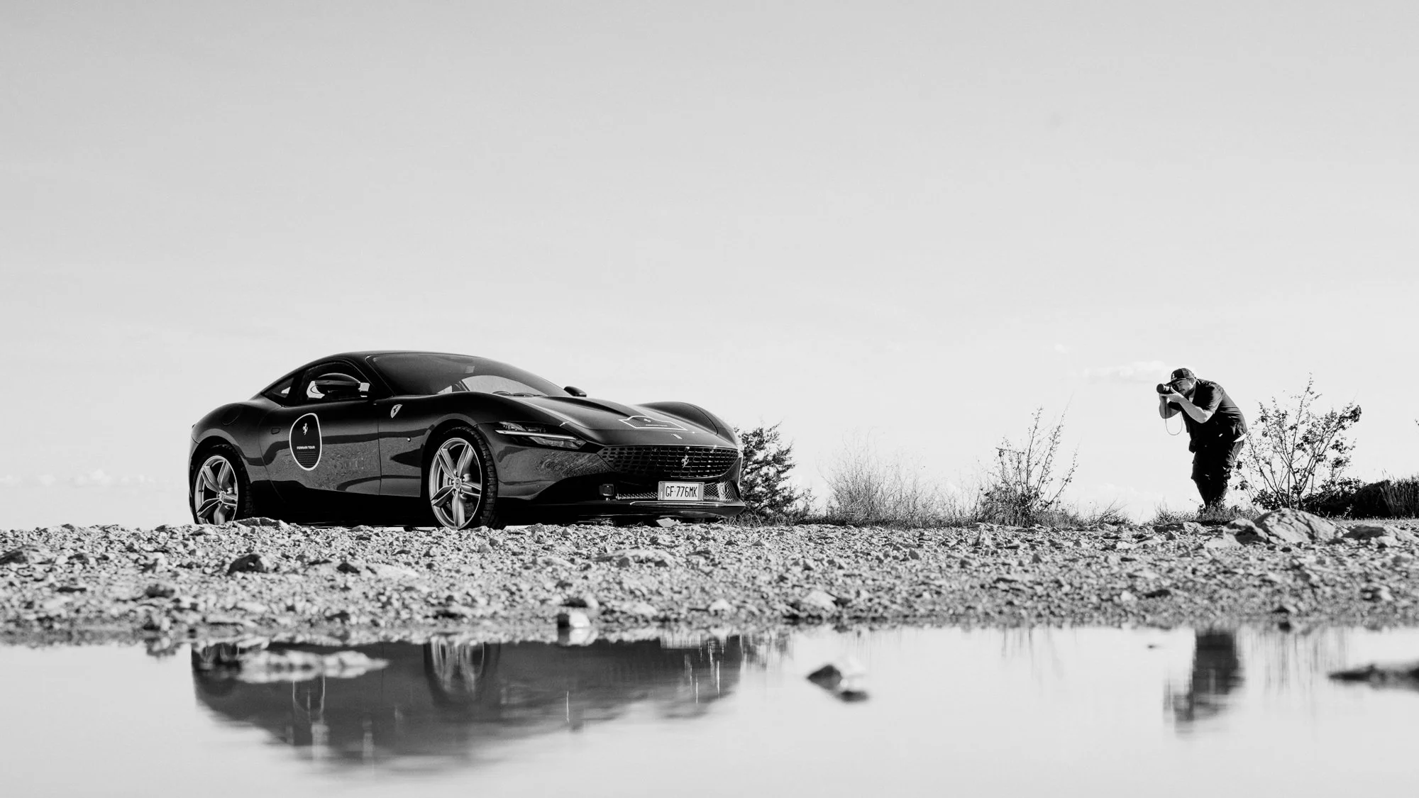 Une voiture Ferrari Roma stationnée sur un terrain rocheux avec une personne prenant une photo de près dans un espace extérieur clair et minimaliste.