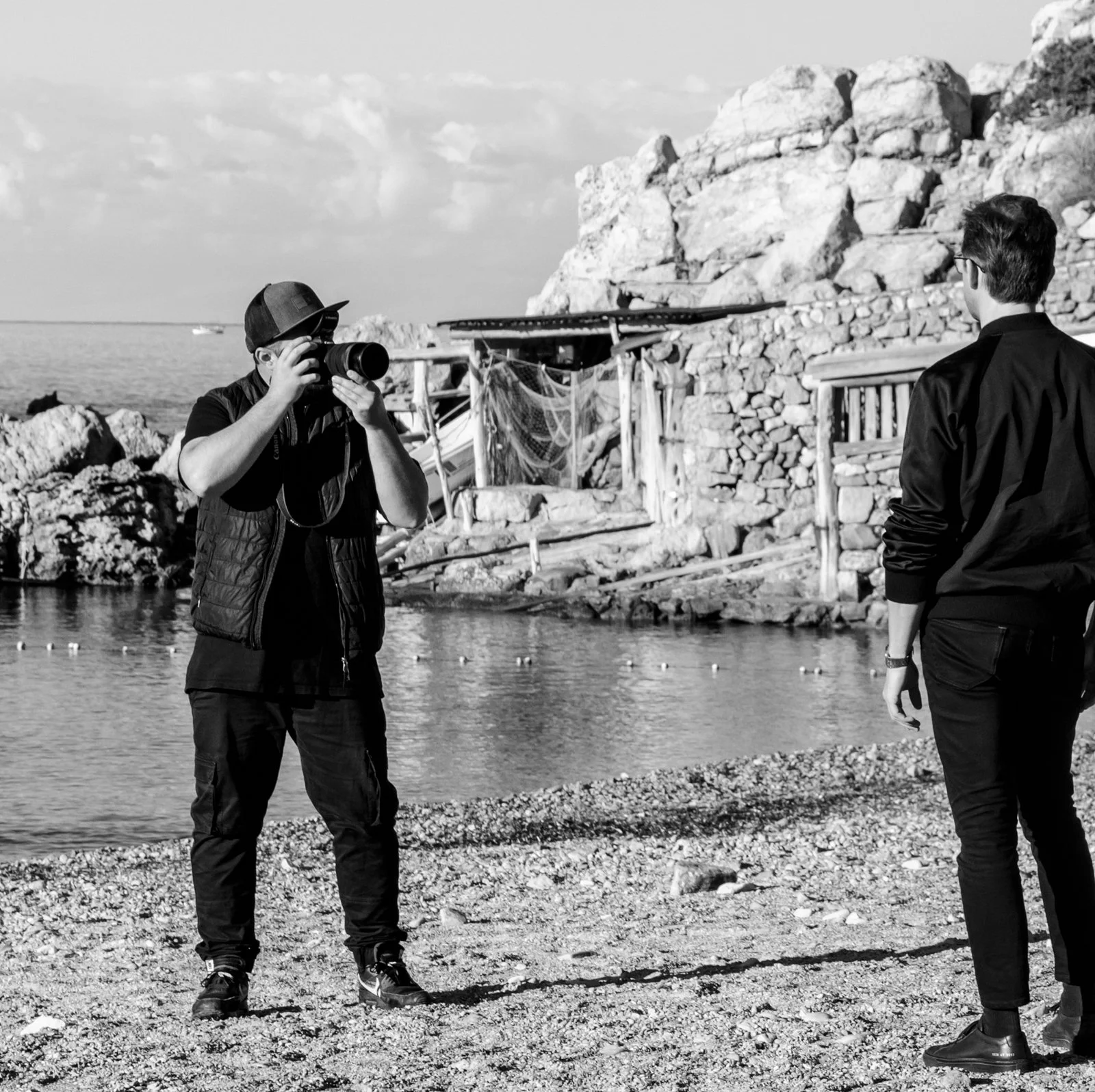 Deux hommes, l'un avec un appareil photo, en train de prendre une photo sur une plage rocheuse avec un rocher et une petite structure en pierre en arrière-plan.