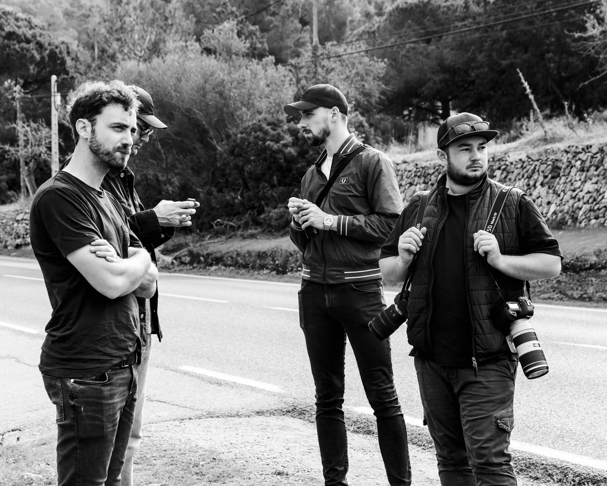 Quatre hommes avec des appareils photo et des sacs à dos sur le bord d'une route, avec un arrière-plan de nature.
