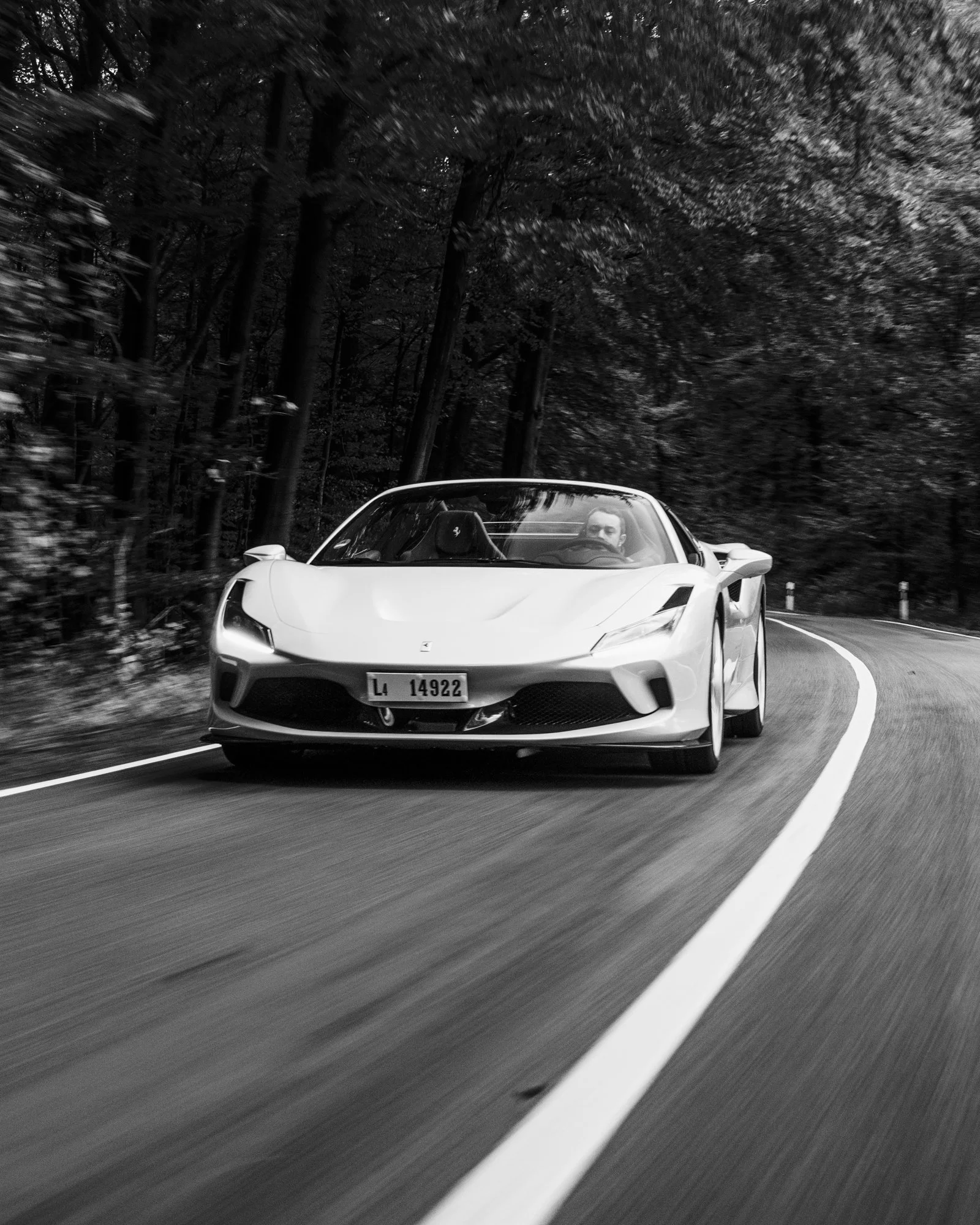 Une voiture Ferrari F8 Tributo conduit sur une route sinueuse entourée d'arbres, dans un environnement forestier, en noir et blanc.