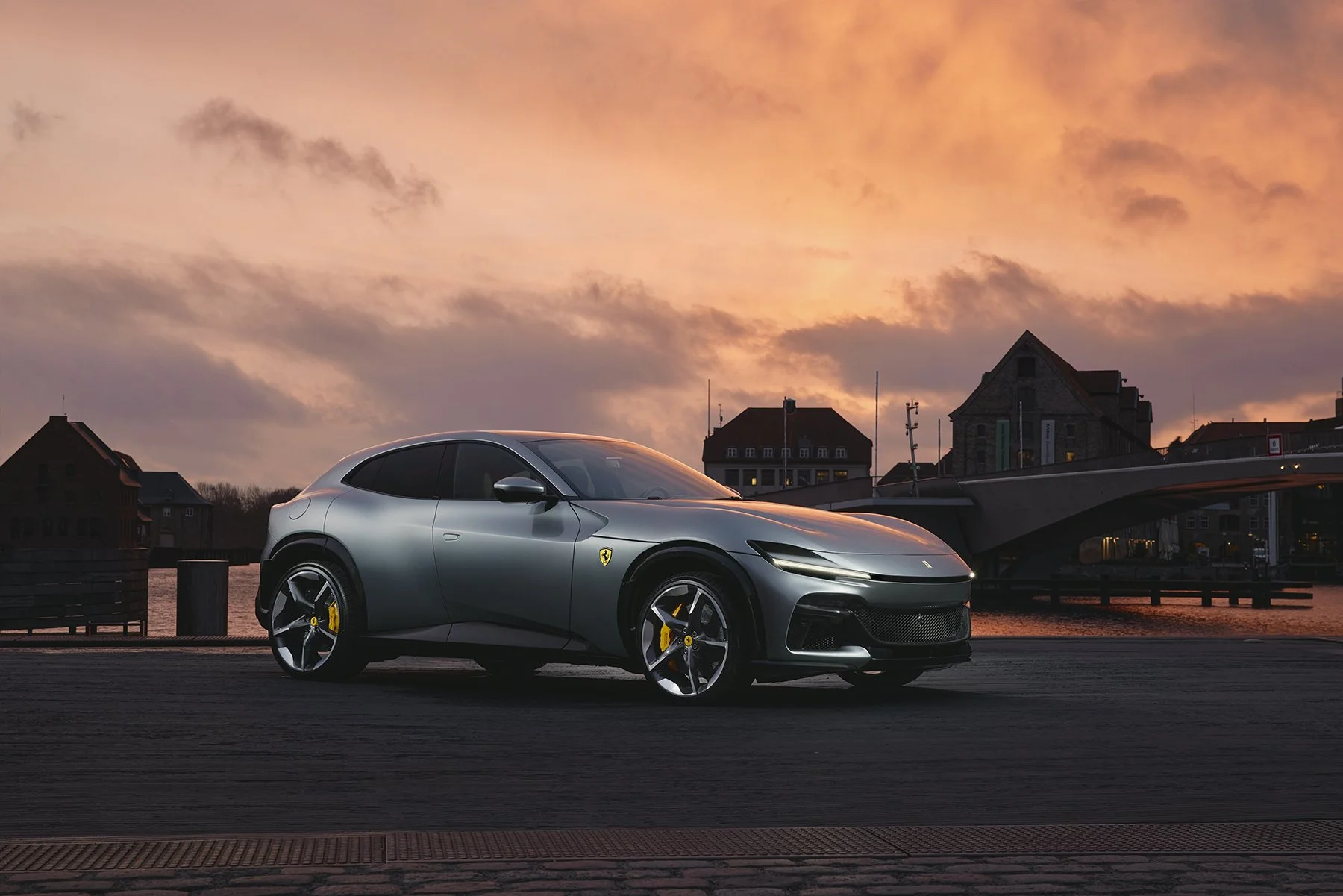 Voiture sportive grise, Ferrari, stationnée près d'un fleuve au lever du soleil, avec un pont et des bâtiments en arrière-plan.