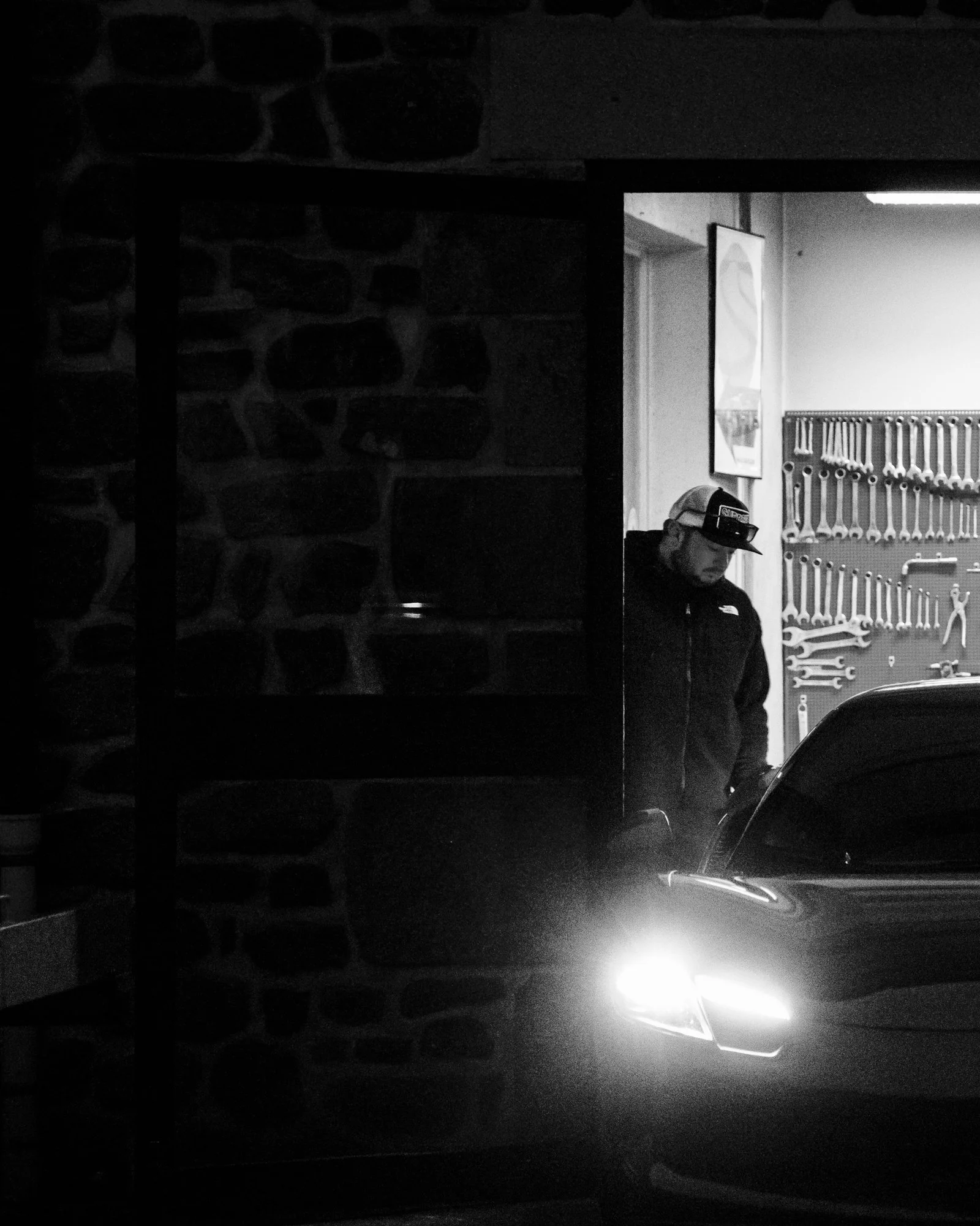 Un homme se tient derrière une voiture éclairée par ses phares, dans un atelier avec des outils muraux.