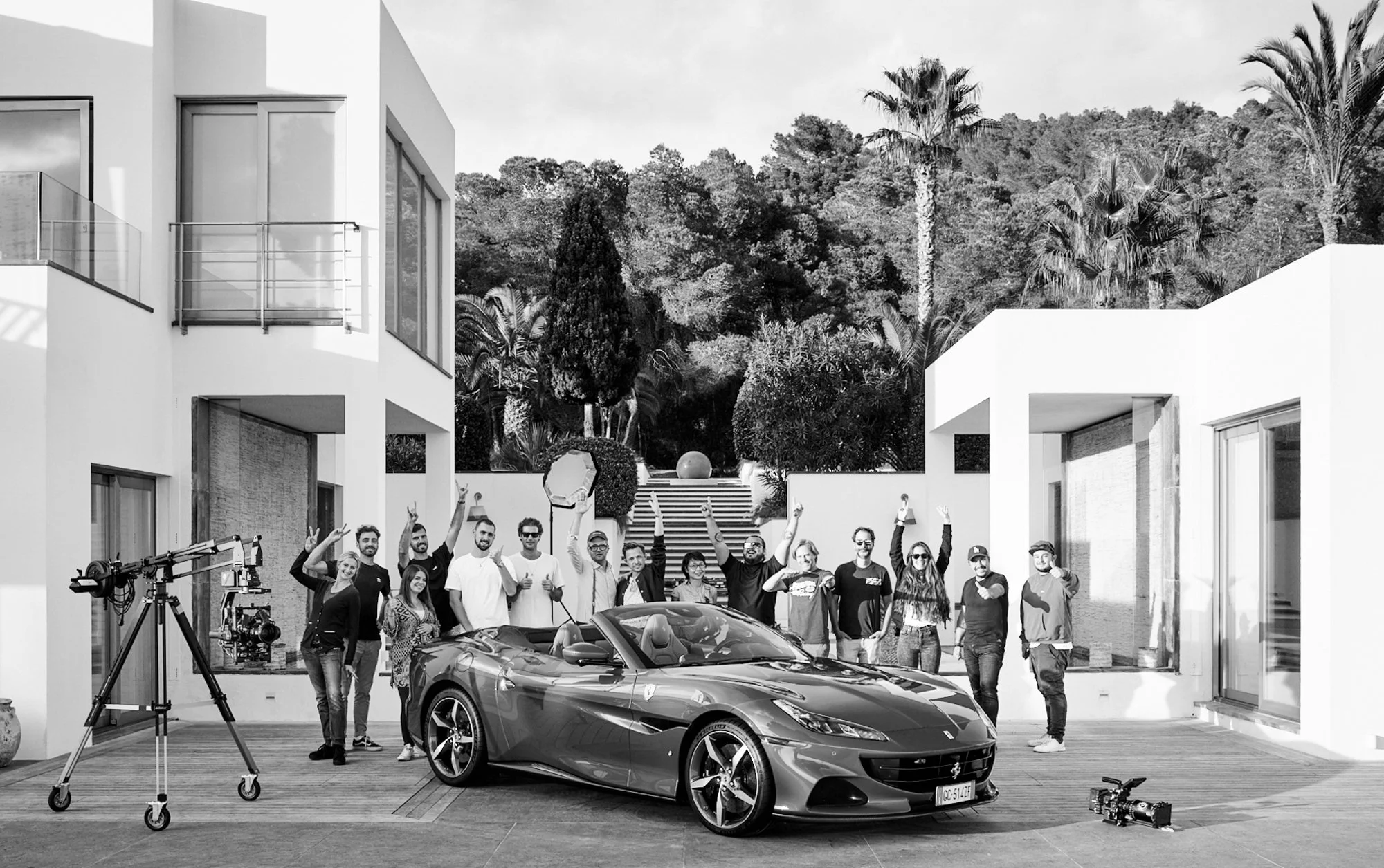 Un groupe de personnes posant devant une voiture de sport décapotable, avec une caméra professionnelle à gauche, dans une résidence moderne entourée de végétation.