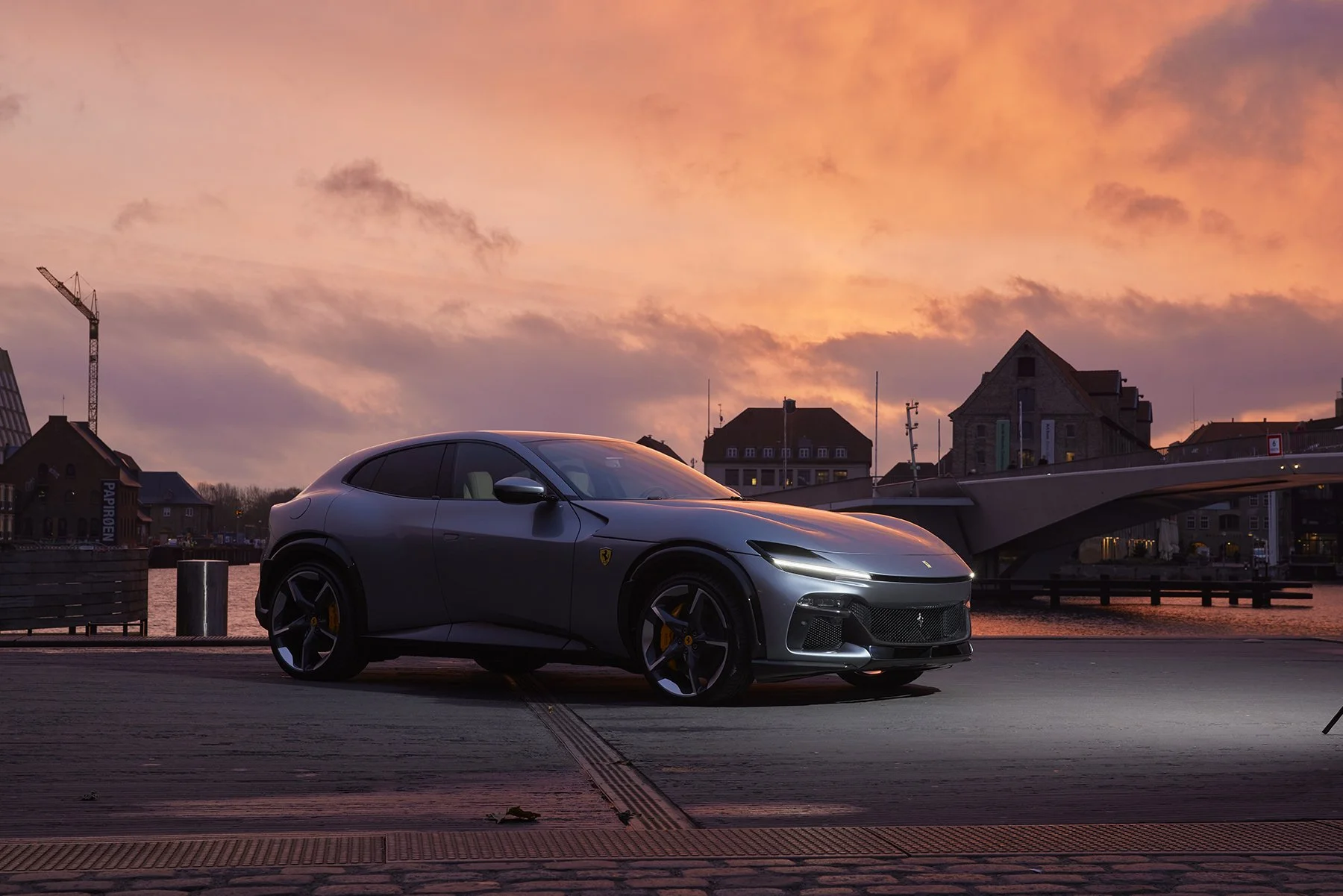 Voiture sportive Ferrari grise garée au bord de l'eau lors d'un lever de soleil à Copenhague, avec un pont moderne en arrière-plan.