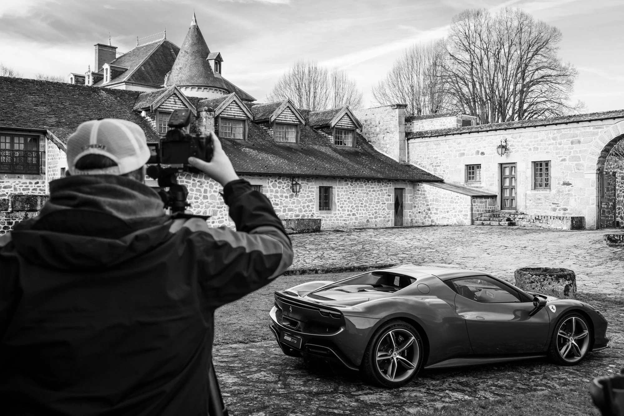 Un homme prend en photo une voiture de luxe Ferrari dans une cour pavée devant un vieux bâtiment en pierre, avec des arbres dénudés en arrière-plan. L'image est en noir et blanc.