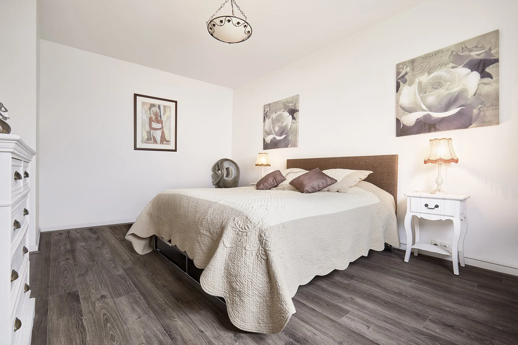 Chambre à coucher avec un lit king-size, draps blancs, oreillers gris, tableaux de fleurs blanches et une lampe sur une table de chevet