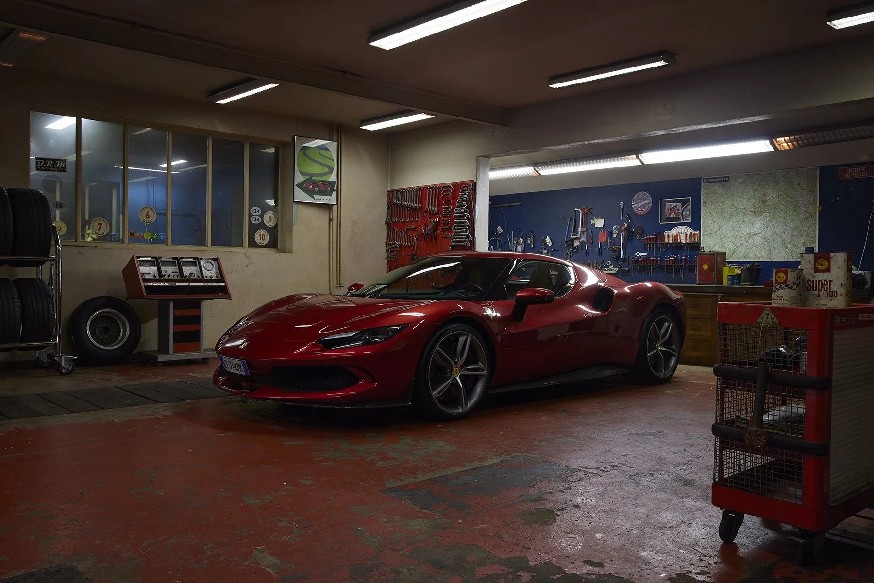 Voiture de sport rouge dans un atelier mécanique avec outils accroché au mur et pneus empilés.