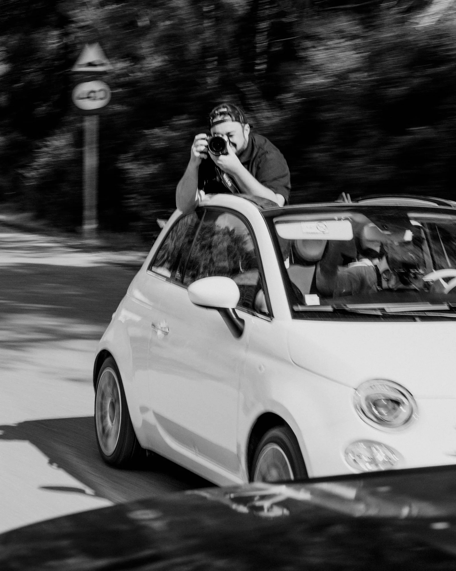 Une personne se tenant à la fenêtre du toit d'une petite voiture blanche en train de prendre une photo avec un appareil photo, dans un environnement urbain en mouvement.