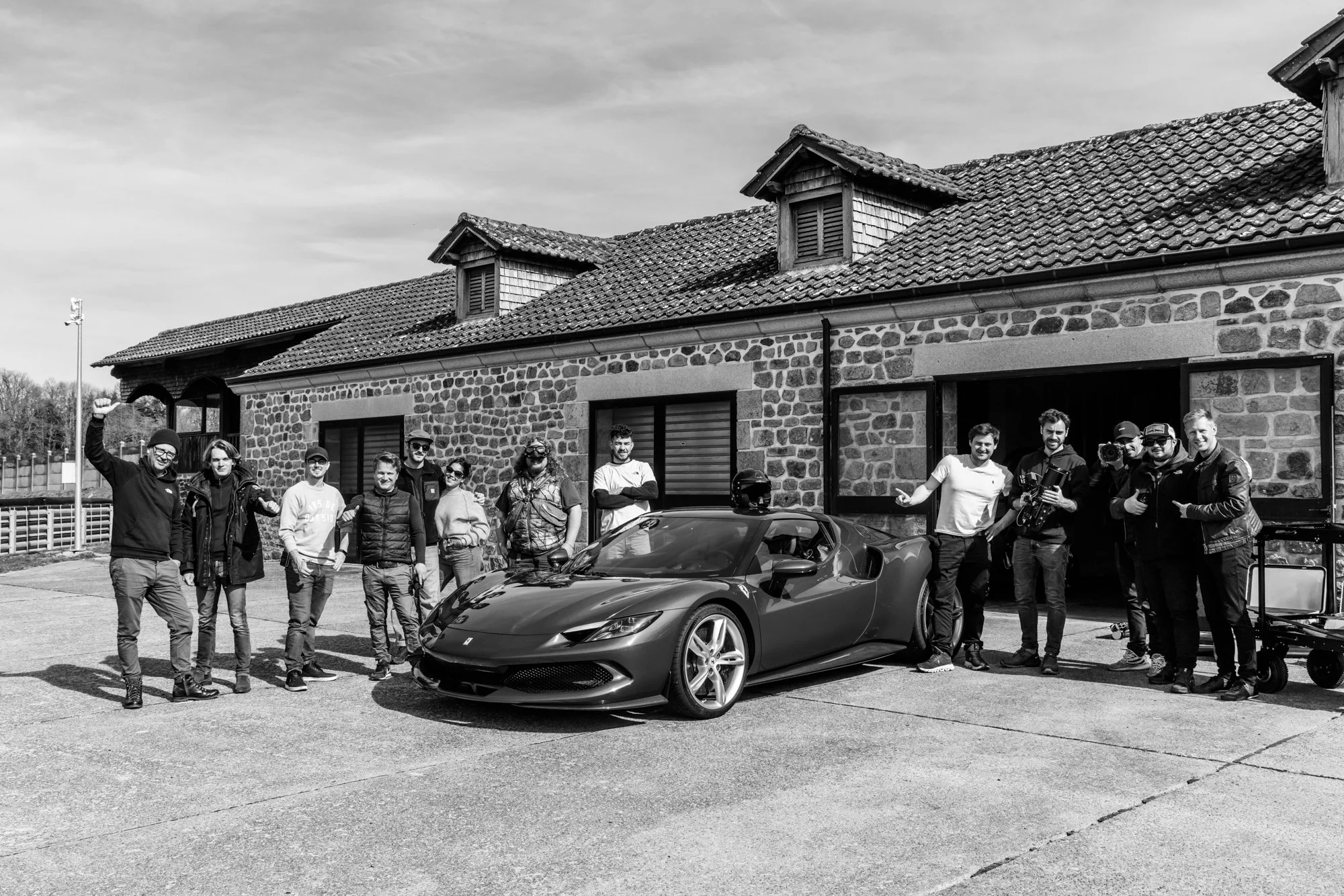 Groupe de personnes posant avec une voiture de sport devant une maison en pierre.