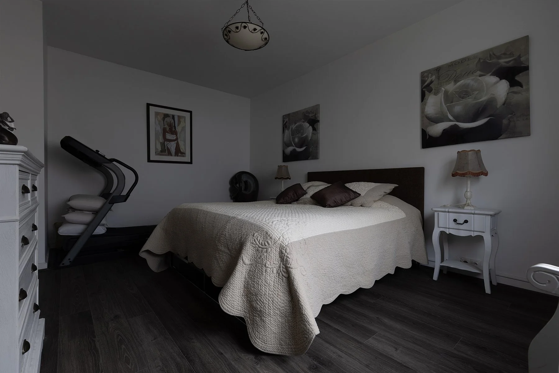 Chambre à coucher avec un lit, des tableaux sur le mur, une lampe de chevet, une machine à tapis roulant dans un coin, et un sol en bois foncé.
