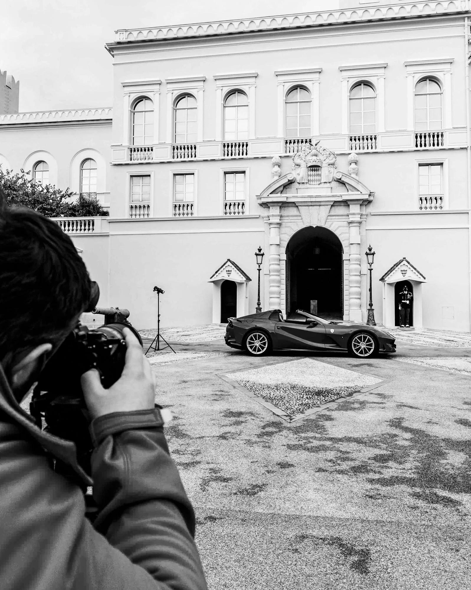 Photographie en noir et blanc d'une voiture Ferrari 812 GTS stationnée devant le Palais de Monaco. Un photographe en avant-plan prend une photo de la voiture.