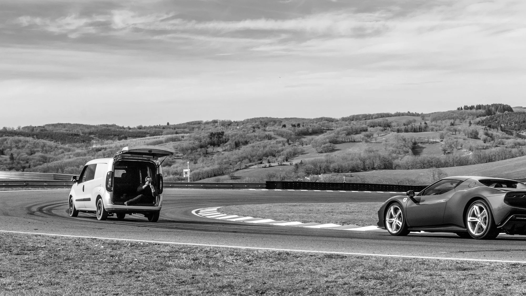 Une voiture de sport Ferrari 296 GTB et un véhicule utilitaire sur une courbe du circuit le Mas du Clos avec un paysage de collines en arrière-plan, en noir et blanc.