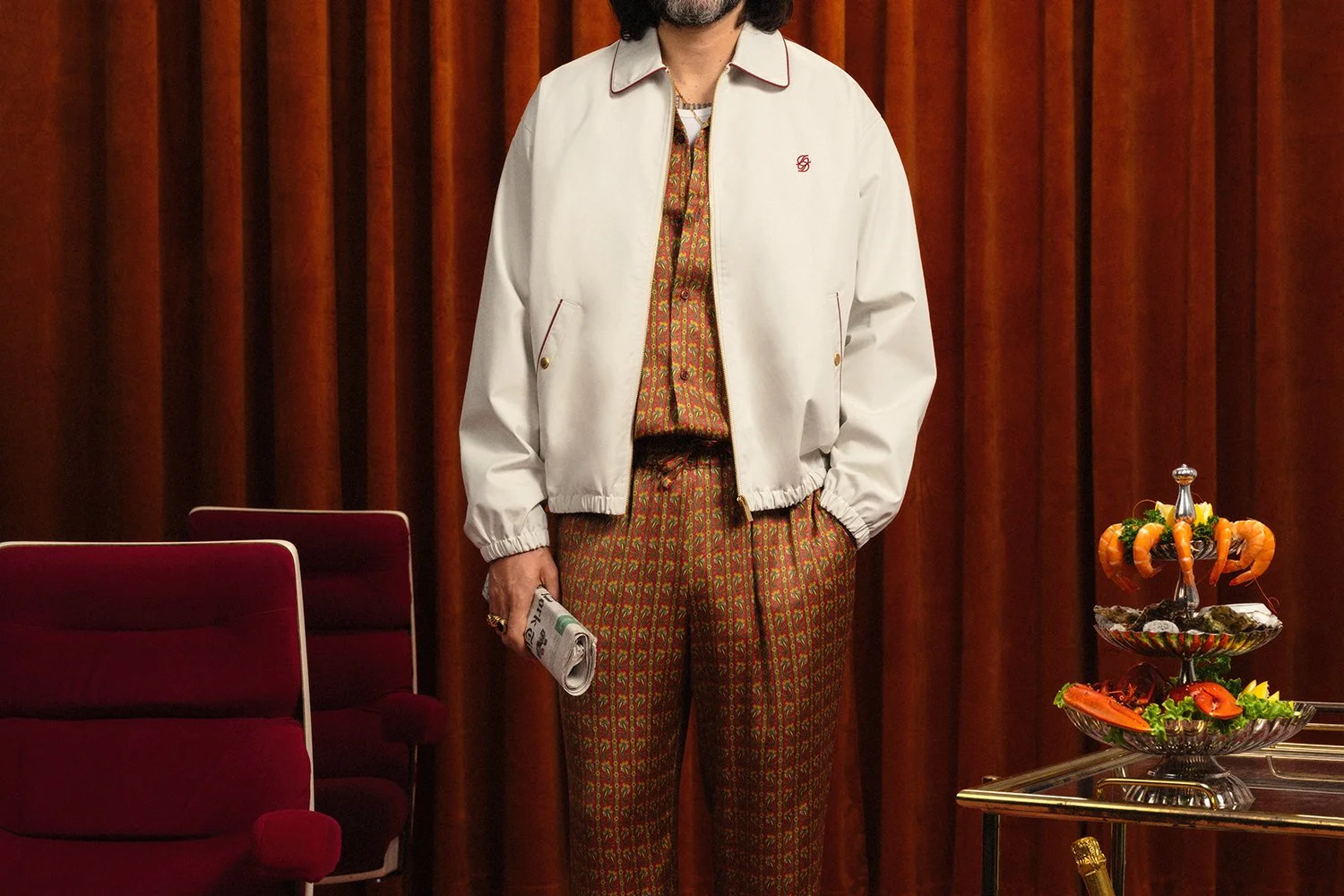 Personne portant une veste blanche, un pantalon à motif rouge et or, tenant un journal, dans une pièce avec des rideaux et un plateau décoratif.