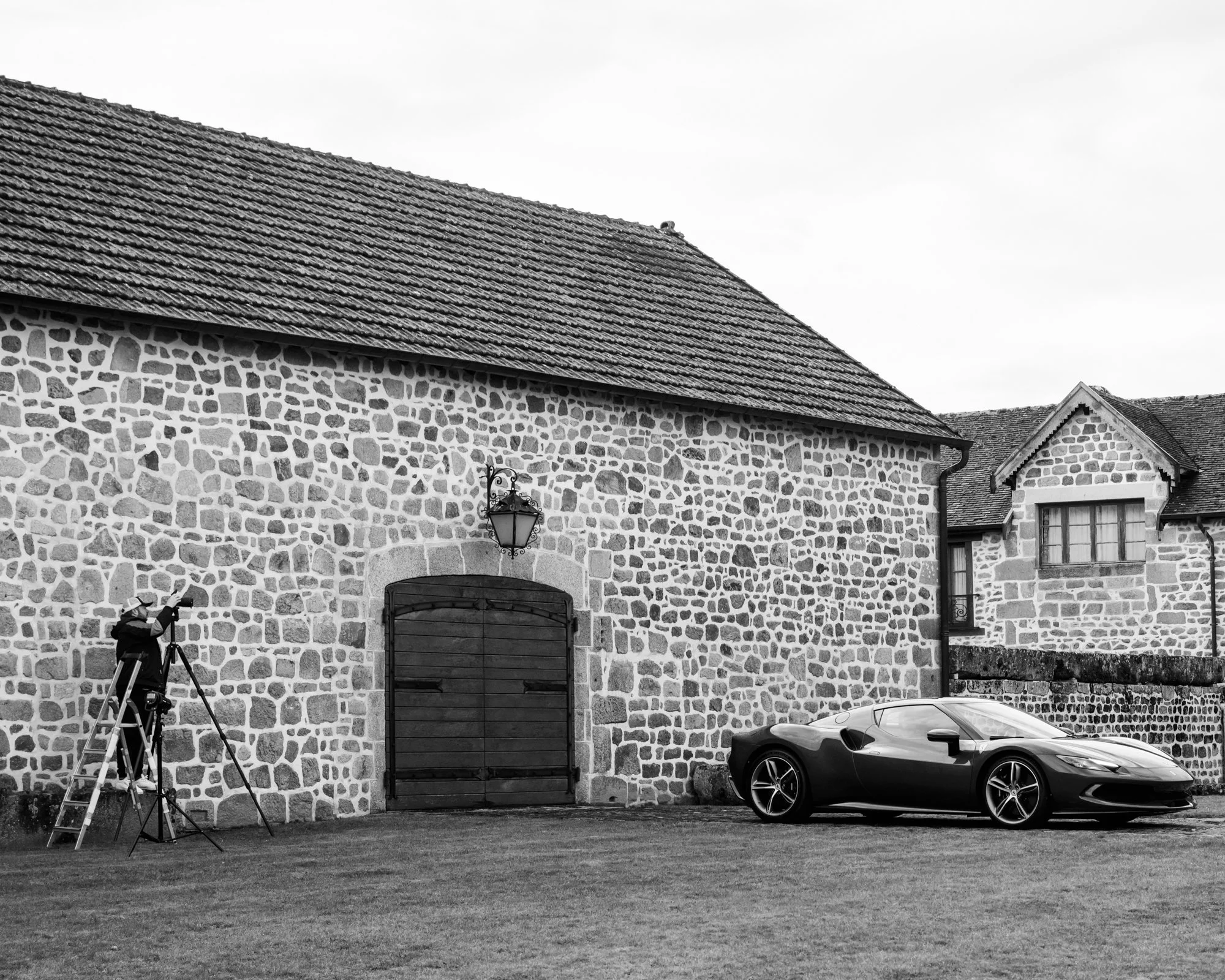 Voiture de sport garée devant un bâtiment en pierre avec toit en tuiles. Personne avec un appareil photo sur un escabeau à gauche.