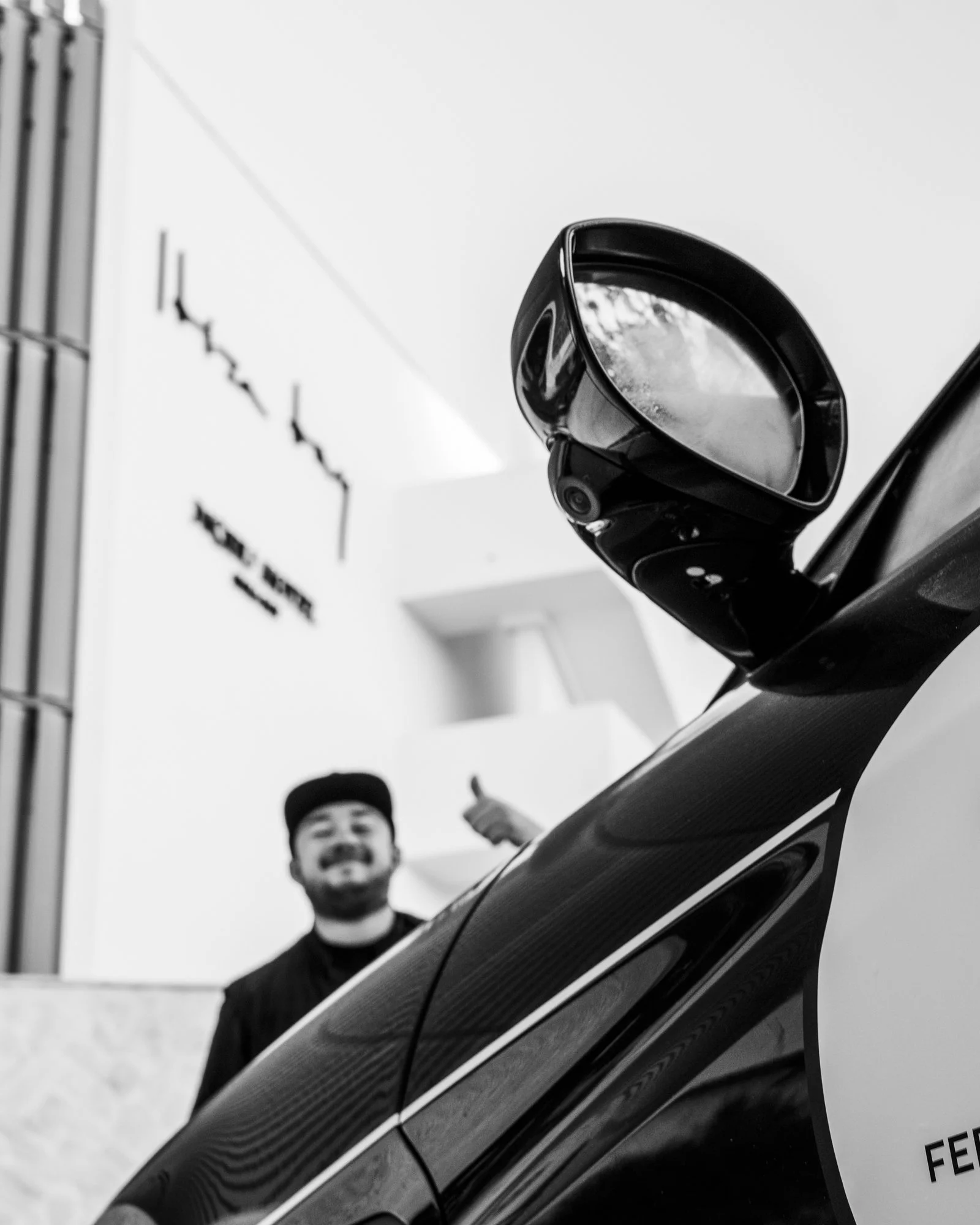 Photo en noir et blanc d'un rétroviseur de voiture de luxe, un homme souriant en arrière-plan faisant un signe de pouce en l'air, dans un intérieur moderne