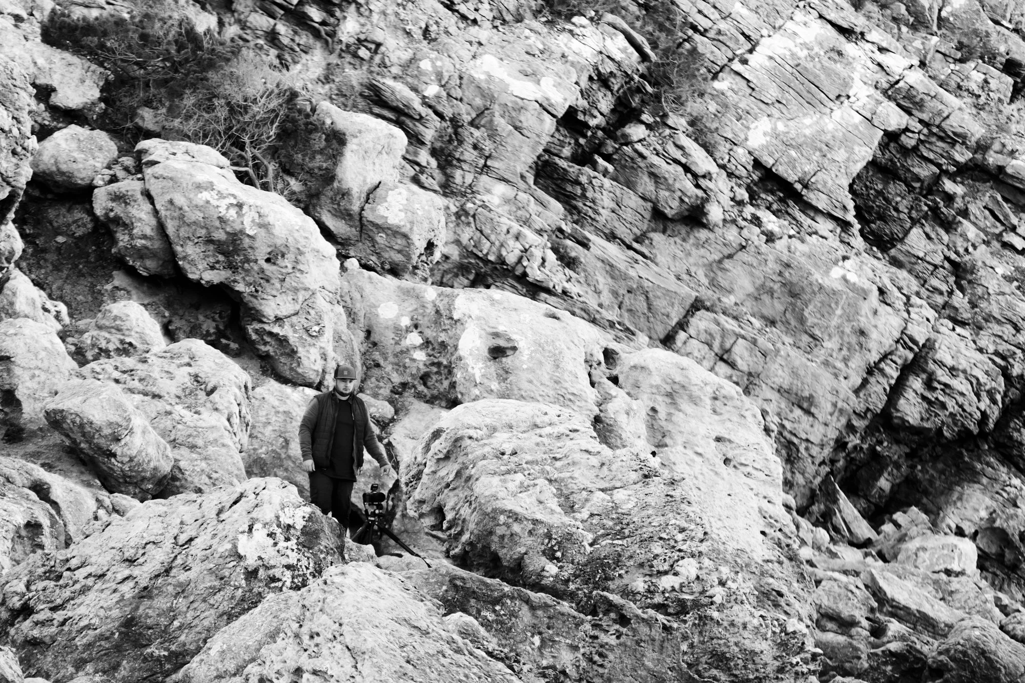 Un homme portant se tient sur un terrain rocheux avec un trépied et une caméra.