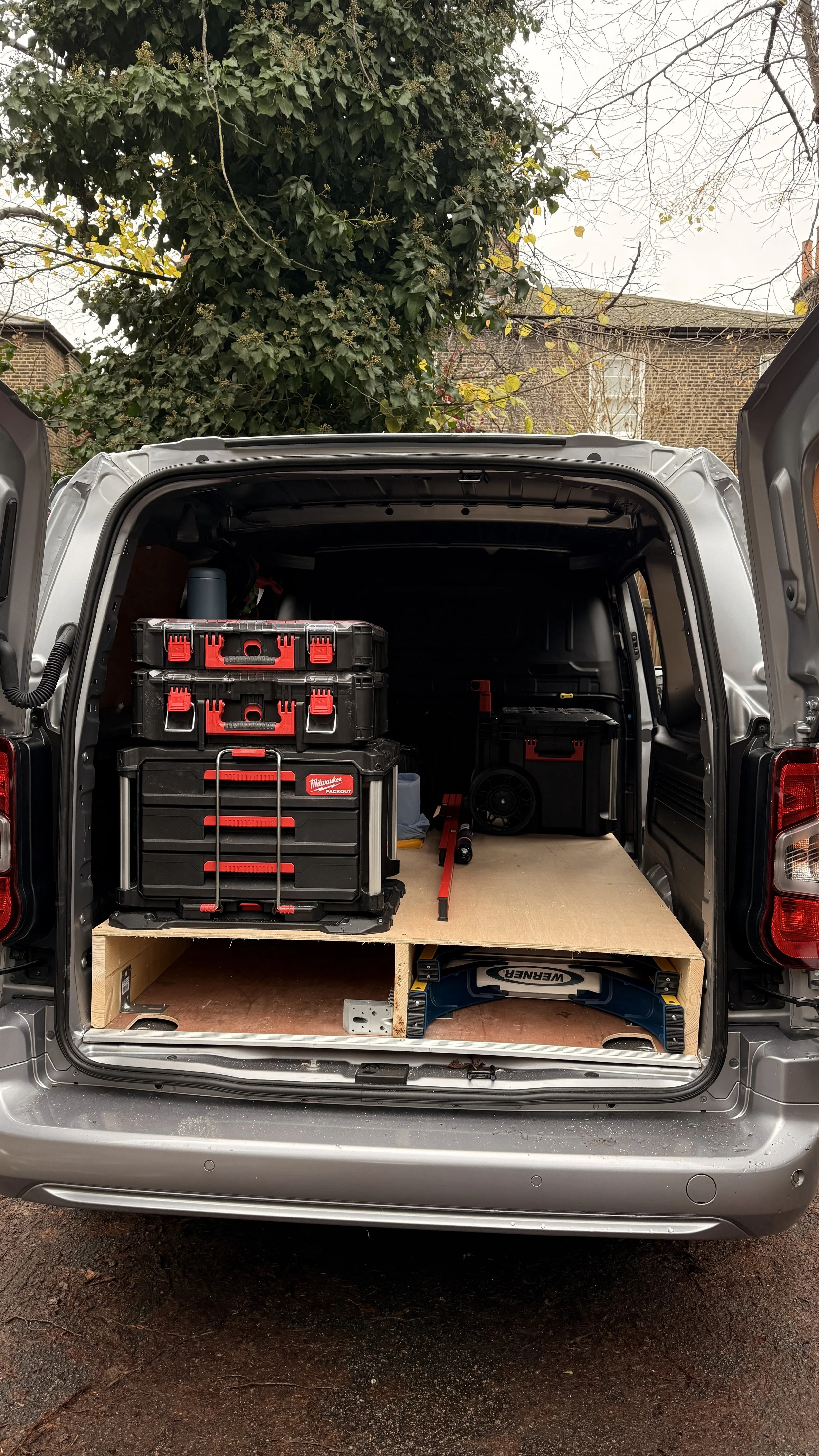 Back of a van with storage boxes, power tools, and a wooden platform for a mobile workshop setup.