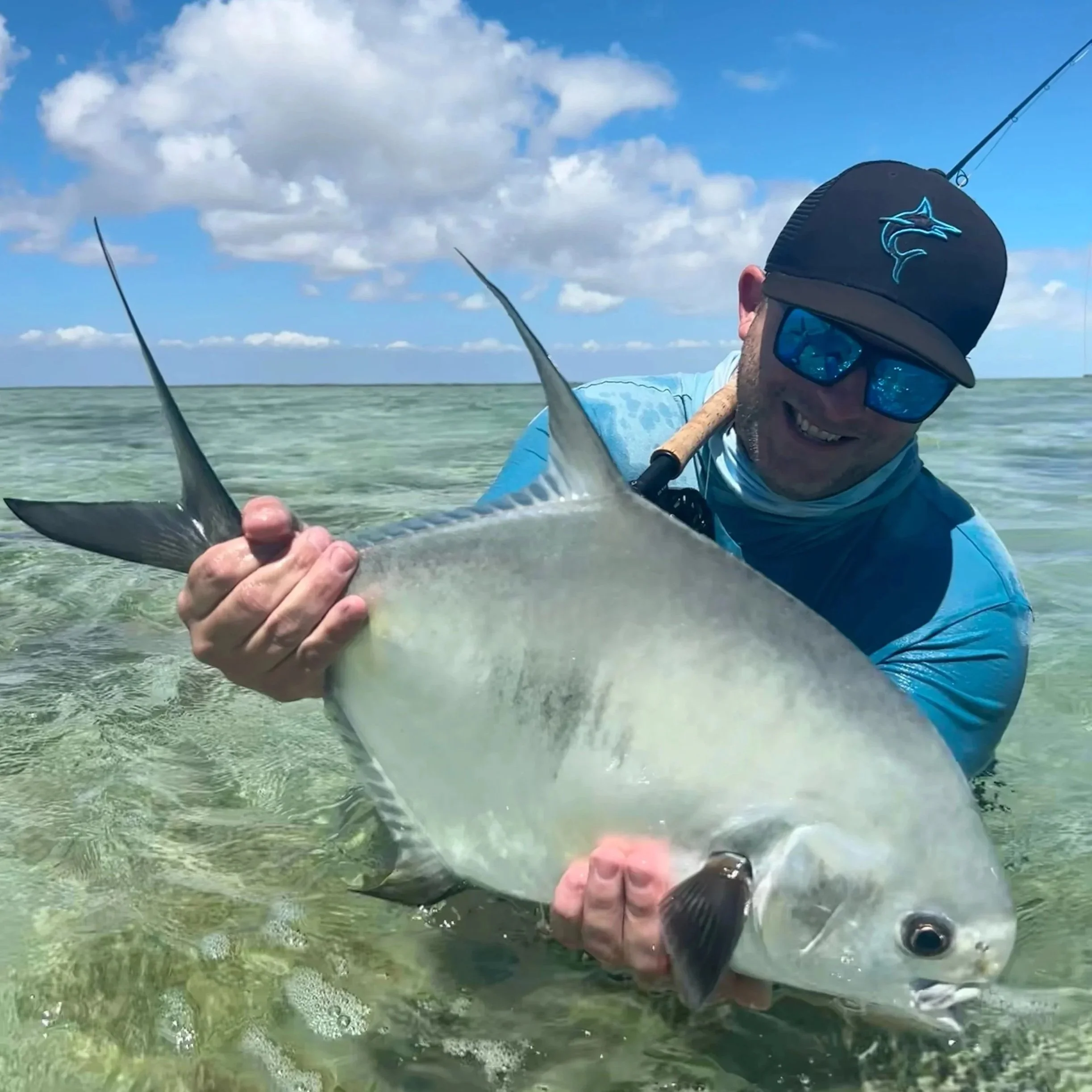 Man in blue shirt and black cap with a fishing logo holding a large fish in shallow water under a partly cloudy sky.