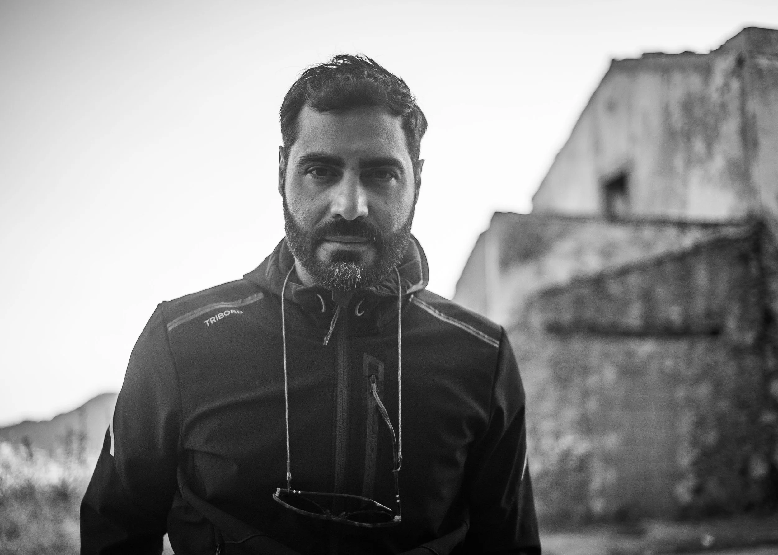 Ritratto di un uomo con barba e capelli corti, indossando una giacca sportiva, all'aperto con sfondo di edifici vecchi e natura, in bianco e nero.