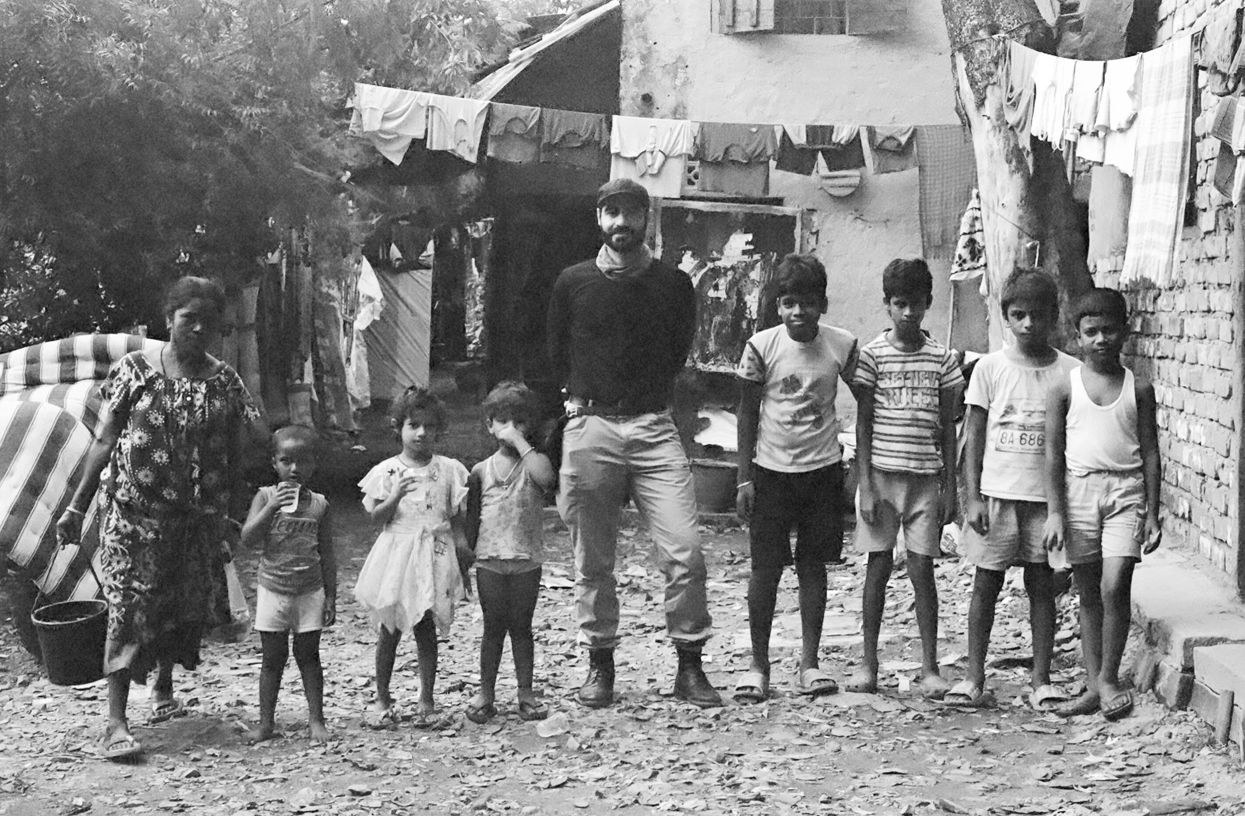 Gruppo di bambini e un adulto in un cortile con case di mattoni, alcune persone stanno facendo il bagno, lavanderia con panni stesi e piante sullo sfondo.