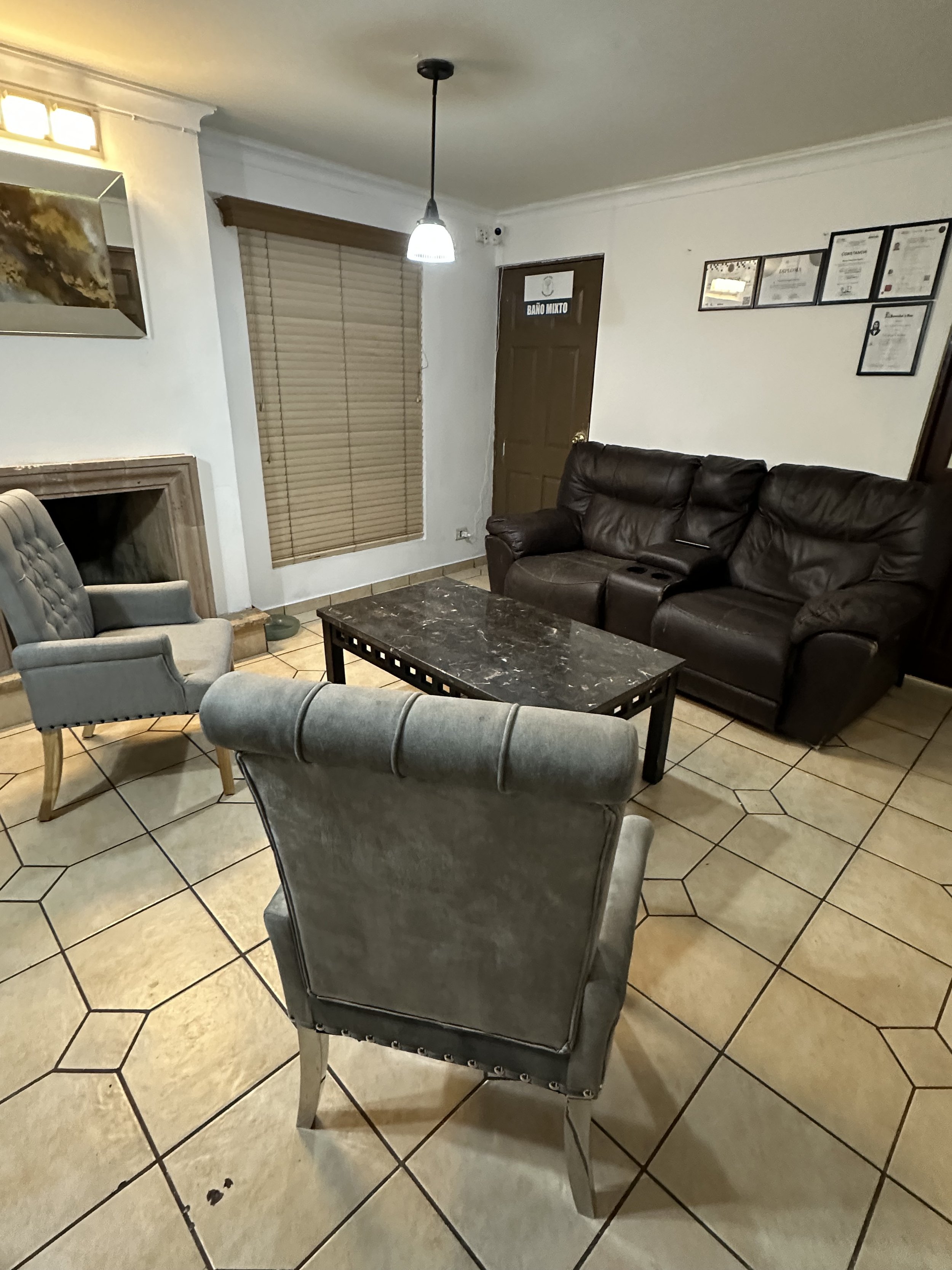 Sala de espera con sillones y una mesa de café en un ambiente decorado con cuadros en la pared y cortinas en la ventana, con un letrero que dice 'Baño Mixto' en la puerta de fondo.