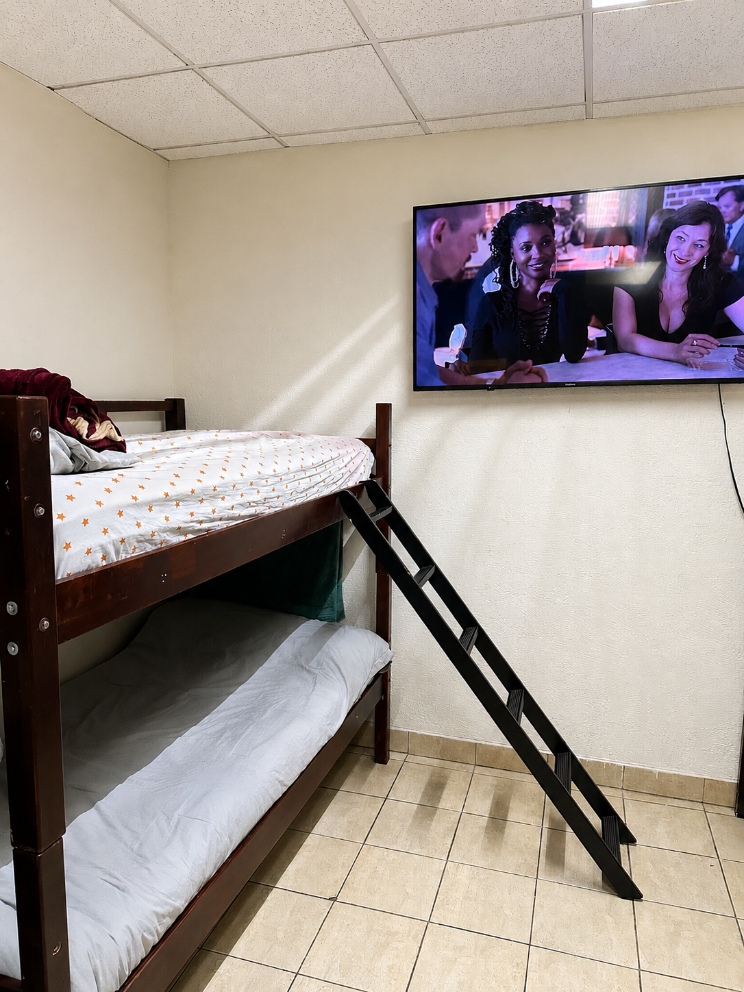Habitación con cama superior y inferior, televisión en la pared mostrando una escena con dos mujeres y un hombre en un restaurante.