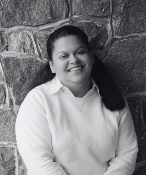 Young woman with dark hair smiling, wearing a light-colored sweater, against a stone wall background.