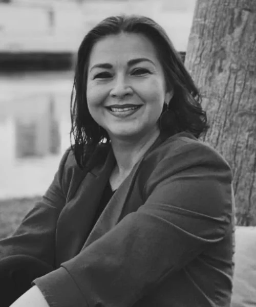 Black and white photo of a woman smiling and sitting outdoors near a tree.