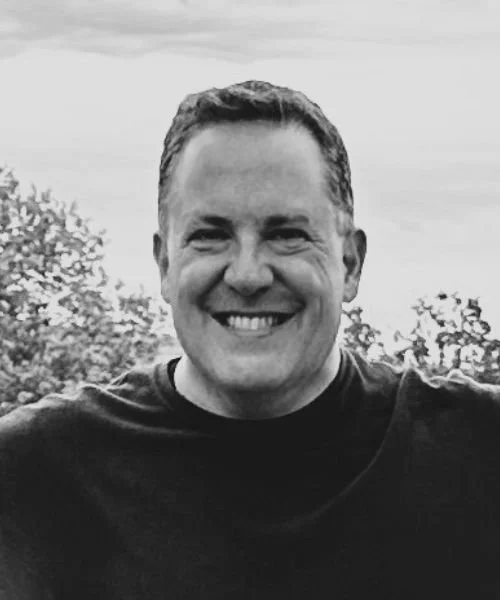 A smiling man with short hair outdoors, with trees and sky in the background, in black and white.