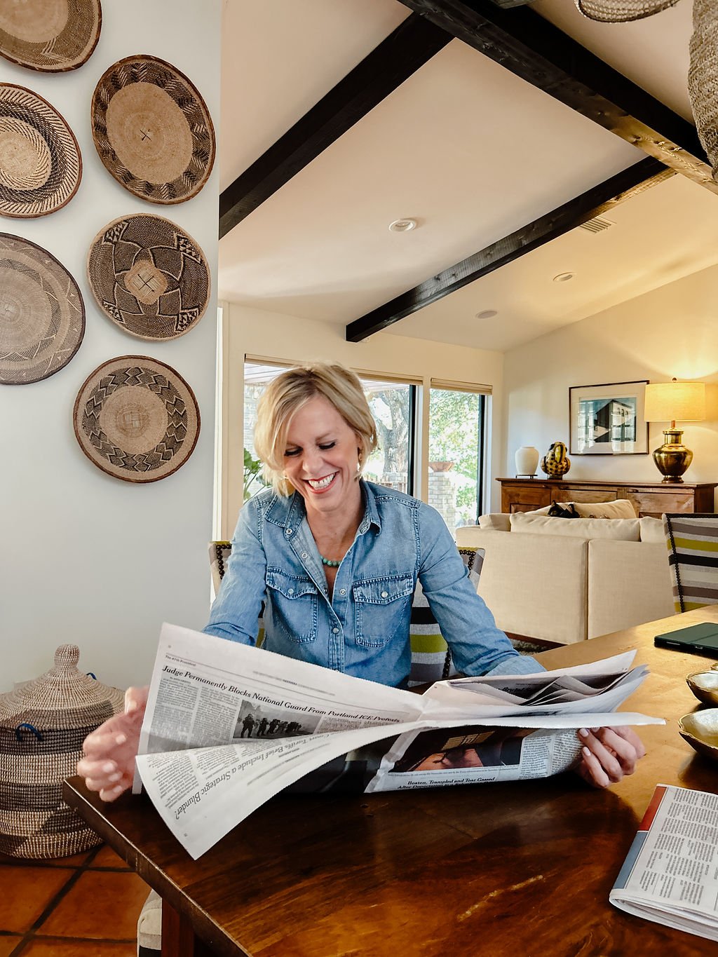 Smiling woman in denim shirt reading a newspaper at a dining table in a well-lit living room with decorative wall hangings, a sideboard, and large windows.