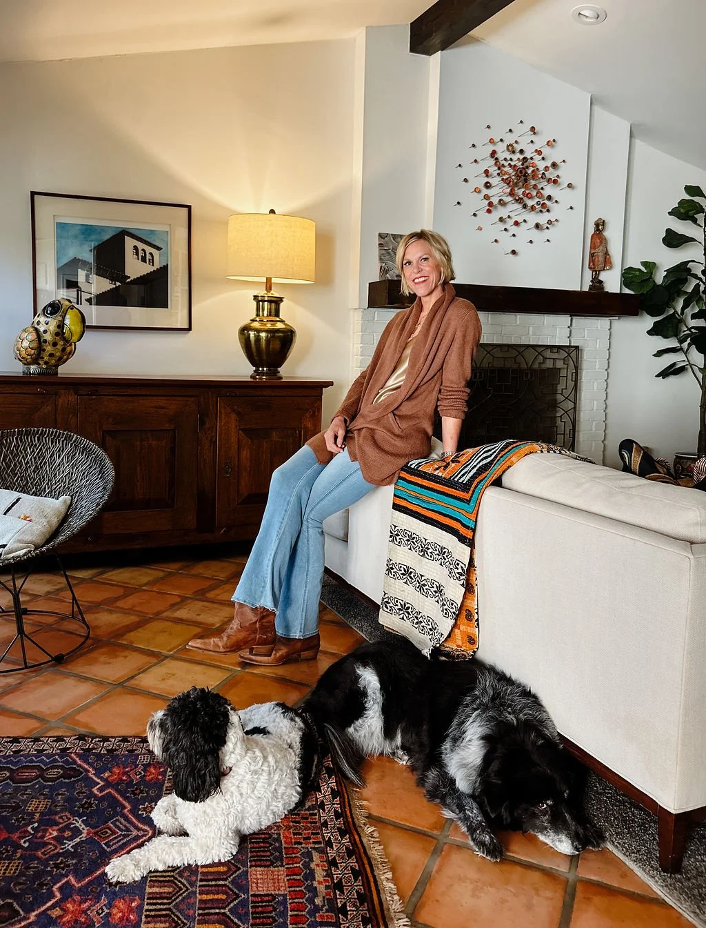 A woman sitting on the arm of a white sofa in a living room, with two dogs lying on the tiled floor in front of her. The room features a wooden cabinet, a yellow lamp, framed artwork on the wall, and decorative items including a metal wall art piece and a sculpture.