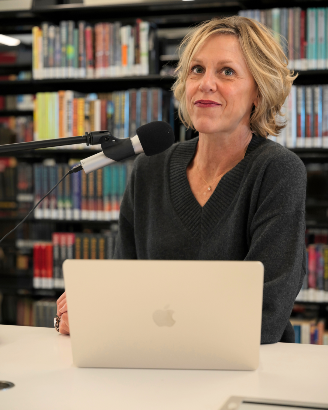 Brooke  Hardie, a woman with blonde hair and a dark sweater sitting at a table with a silver Apple MacBook in front of her, speaking into a black microphone, with a background of bookshelves filled with colorful books.