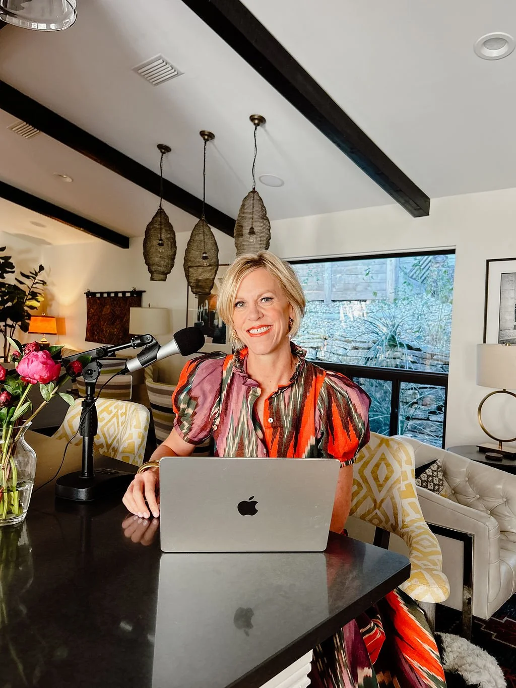 A woman with short blonde hair sitting at a kitchen counter with a MacBook, speaking into a microphone, in a modern living room with decorative lighting, a window with outdoor scenery, and stylish furniture.