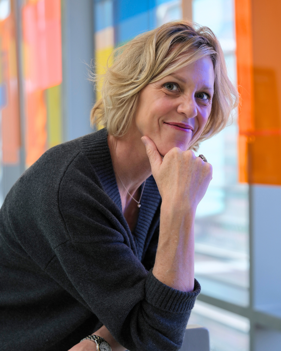Brooke Hardie, esq, with short blonde hair, wearing a dark sweater, sitting in front of a colorful background of blue, yellow, and orange. She is smiling slightly, with her chin resting on her hand, looking at the camera.