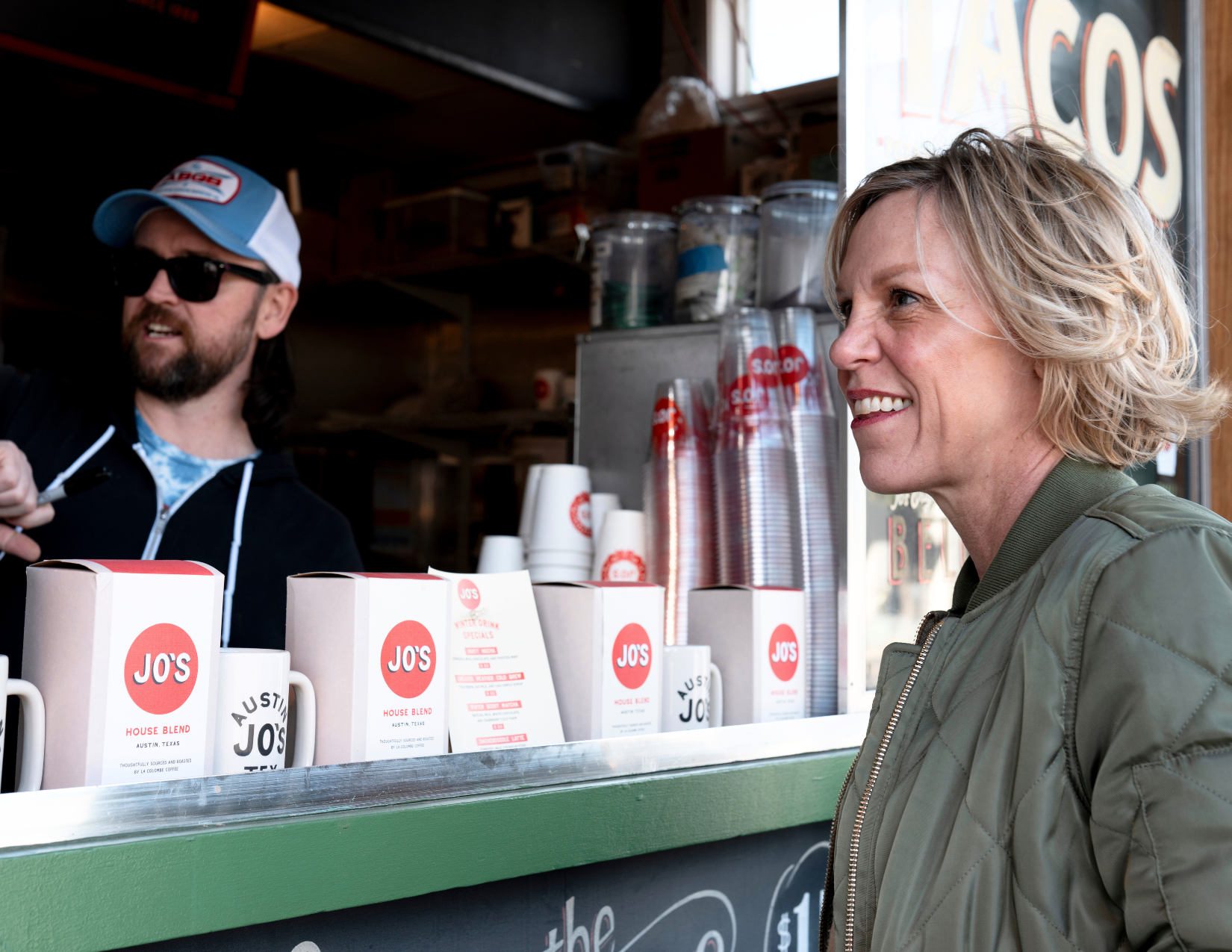 A woman smiling while ordering at a coffee stand, with coffee cups and supplies behind the counter, and a man wearing sunglasses and a hat assisting her.