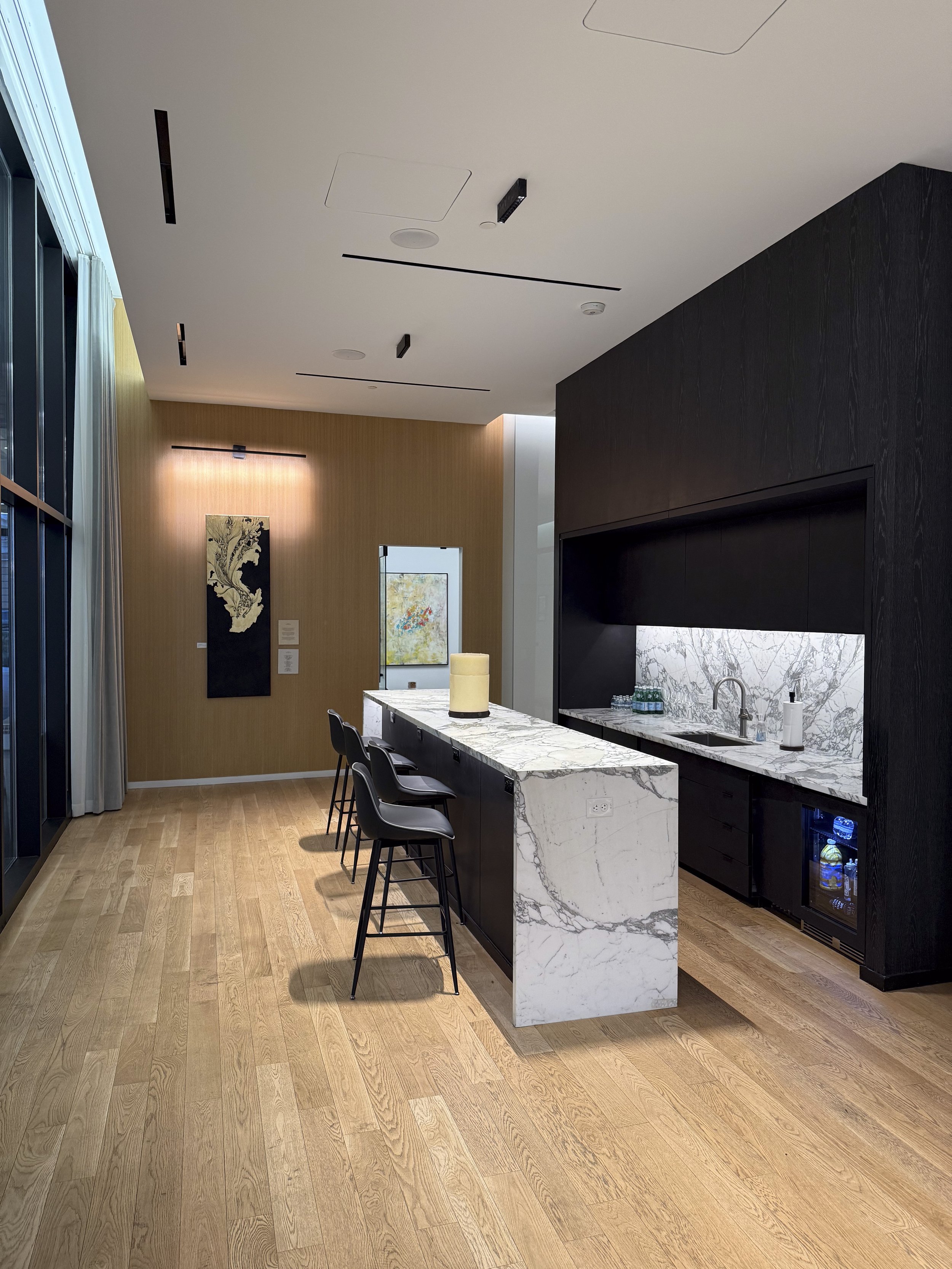 Modern kitchen with a marble island, black cabinets, and a small wine fridge. There are four bar stools at the island and artwork on a wooden wall across from the kitchen.