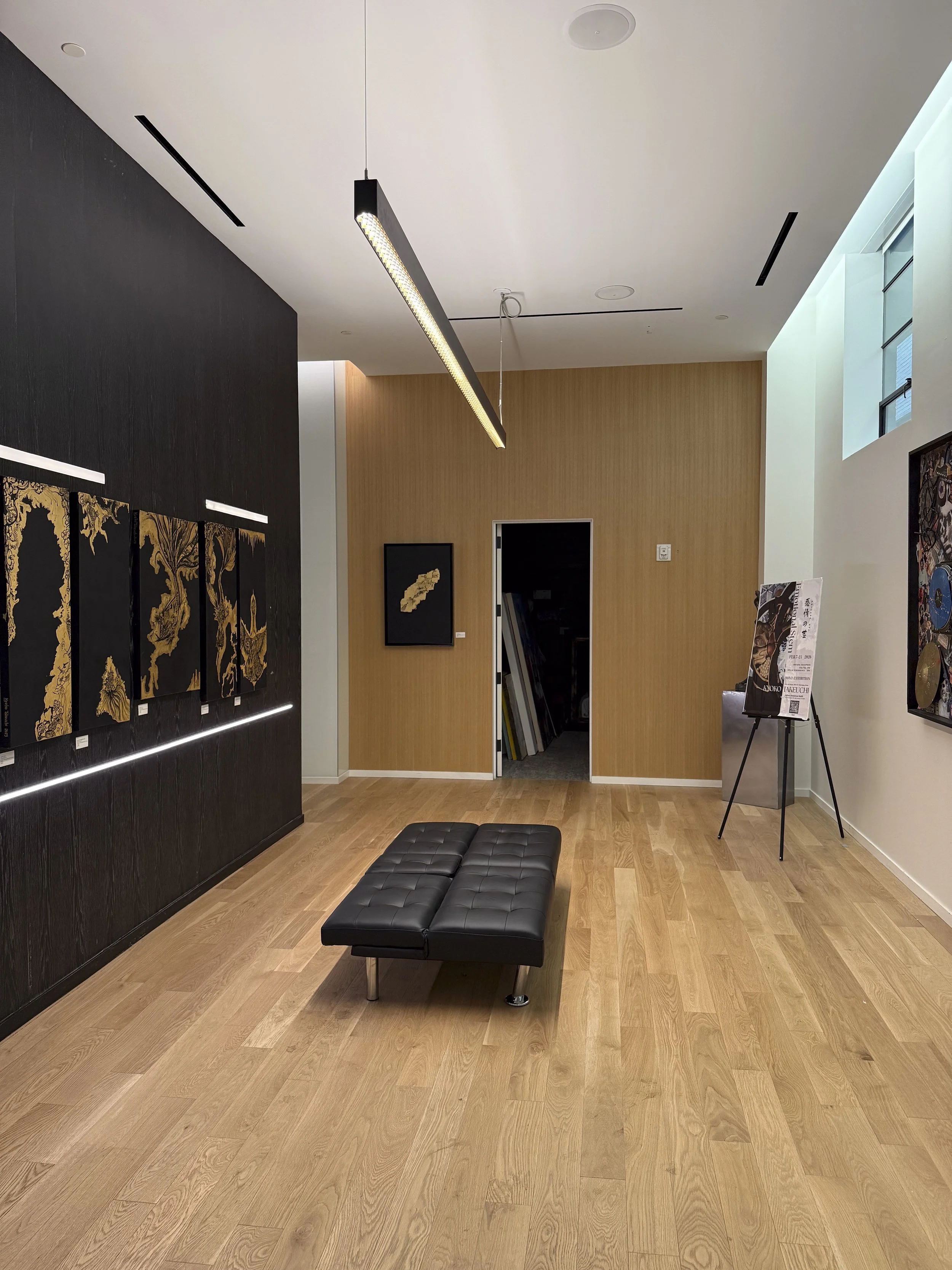 Art gallery interior with black leather bench, artworks on dark and light walls, and a wooden floor.