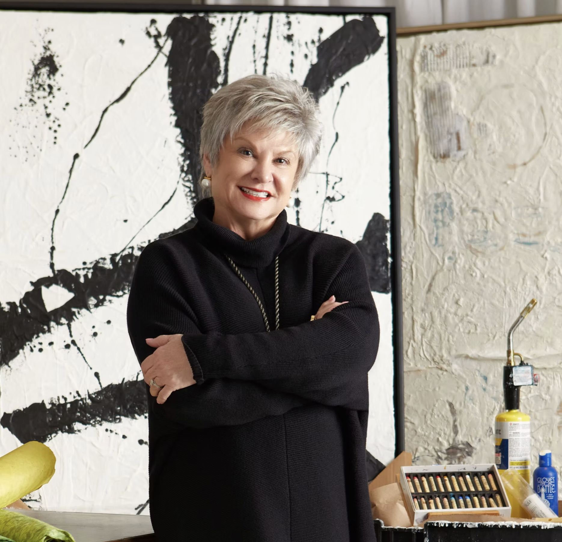Older woman with short gray hair smiling and standing with arms crossed in front of abstract black and white artwork in an art studio or gallery, with art supplies and equipment visible.