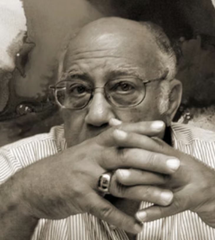 A black and white photo of an elderly man with glasses, bald with some gray hair on the sides, wearing a striped shirt, and resting his face on his clasped hands with a ring on his finger.