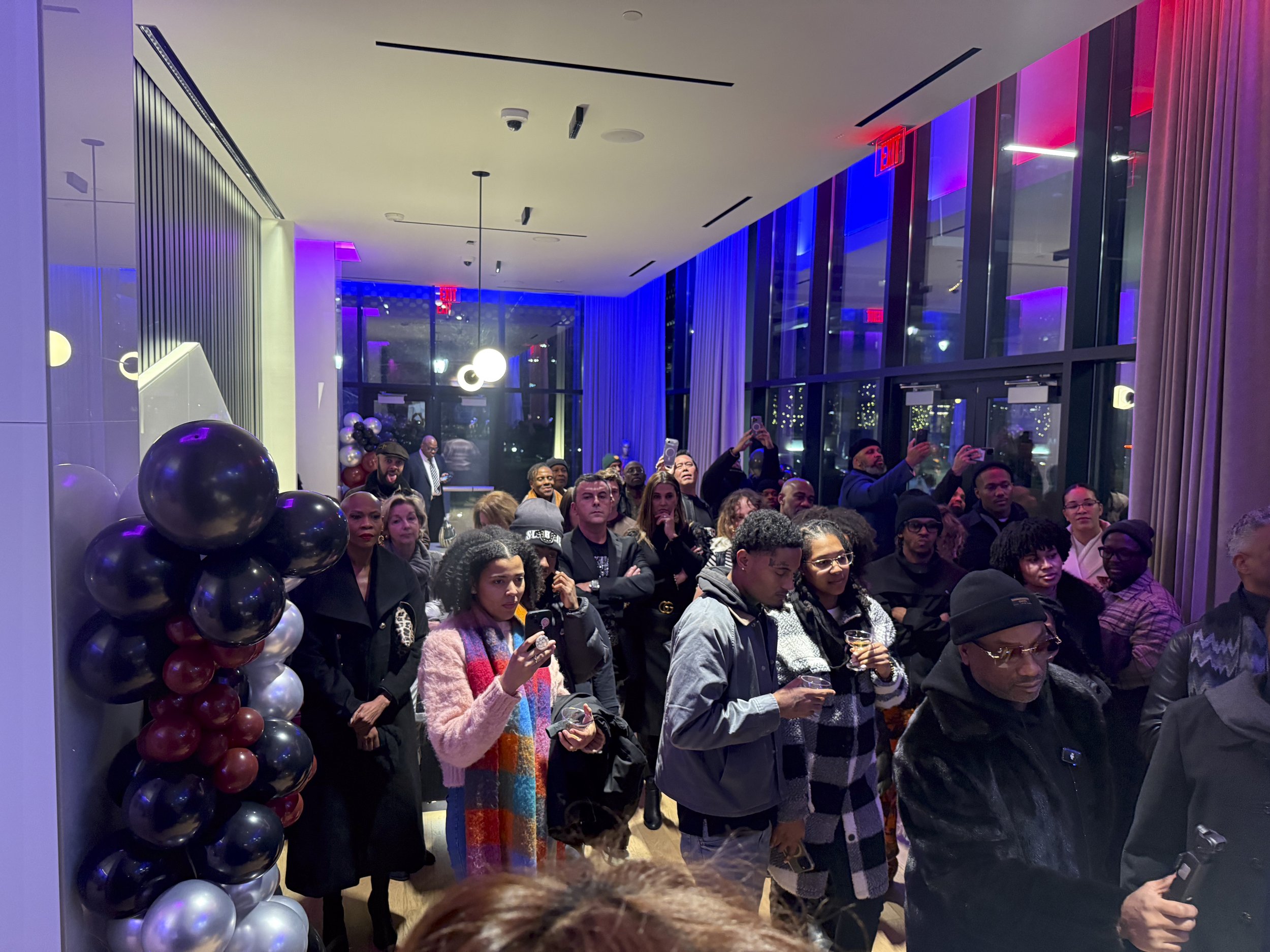 A crowd of people gathered at an indoor event with festive decorations, including balloons in dark shades. The attendees are dressed in stylish clothing, some holding drinks and taking photos, with large windows and colorful lighting in the backgroun