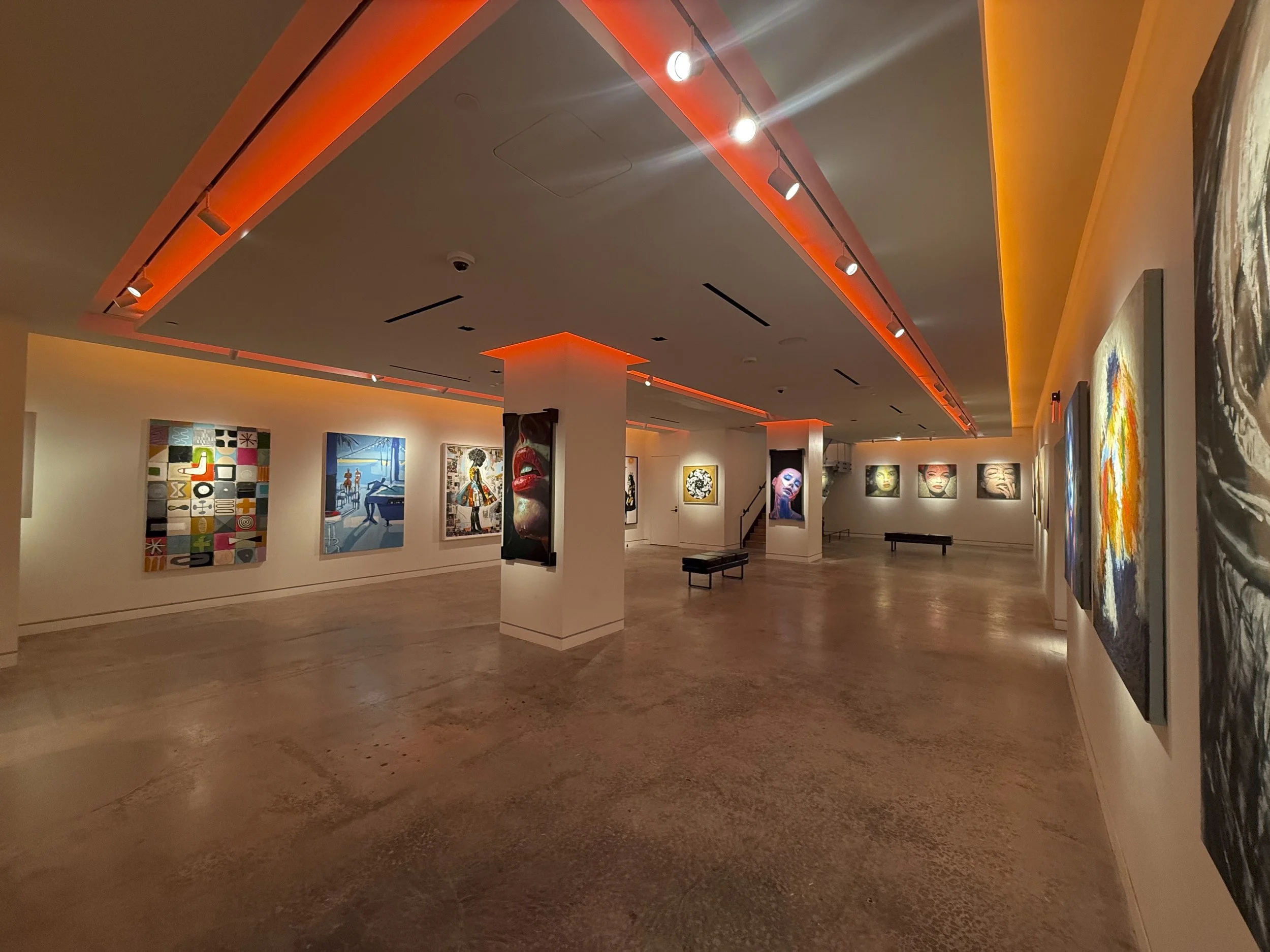 Art gallery with various colorful paintings on white walls, benches, dim lighting with orange accent lights on ceiling.