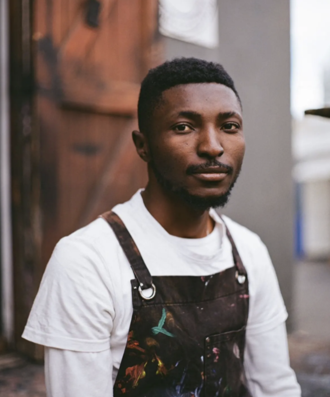 A young man with dark skin, short curly black hair, and a beard, wearing a white t-shirt and a dark apron with paint splatters, standing indoors near a wooden door.