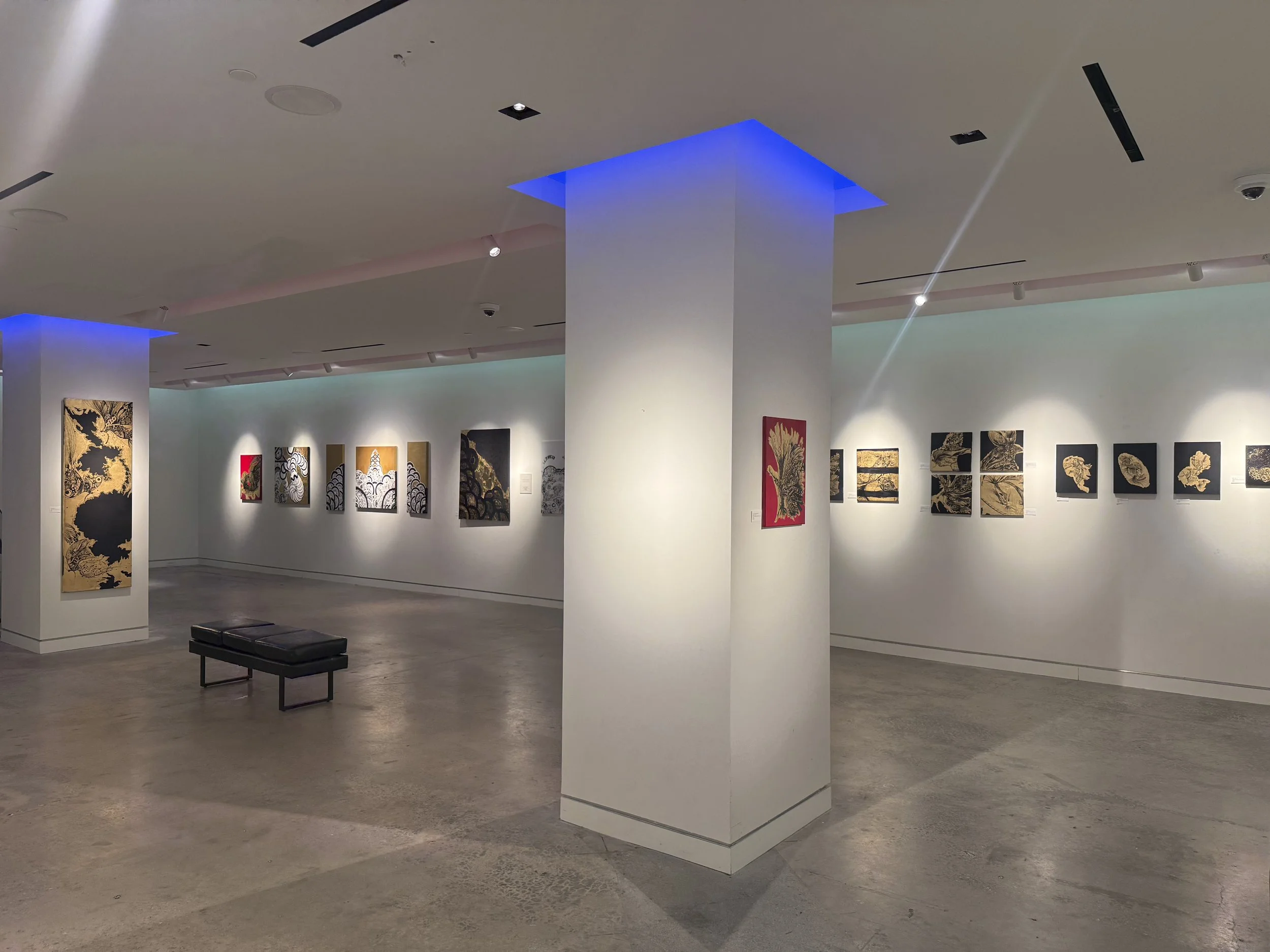 Art gallery with white walls displaying paintings with black and gold patterns, lit by spotlights, and a black bench in the foreground.