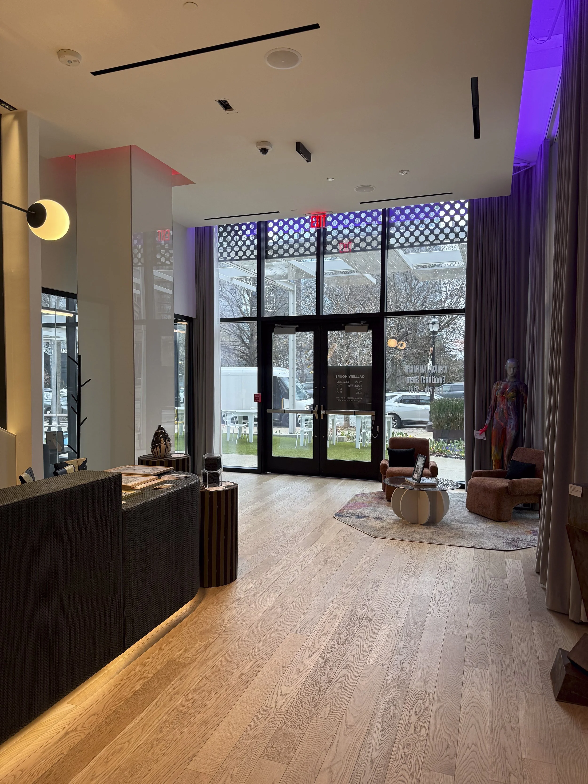 Interior view of a modern lobby or reception area with glass doors, a small sitting area with chairs and a table, and decor elements like a mannequin and artwork.