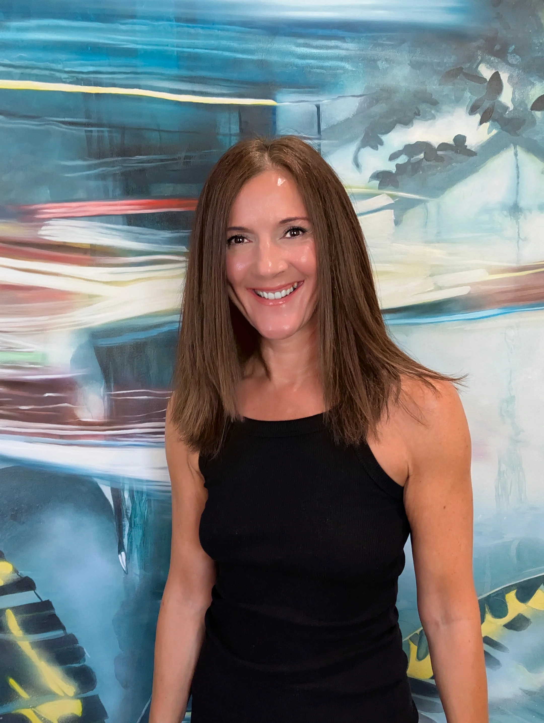 A woman standing in front of a large abstract painting that depicts a forest scene with blurred figures walking through it. She is holding a brush and has a cart with painting supplies.