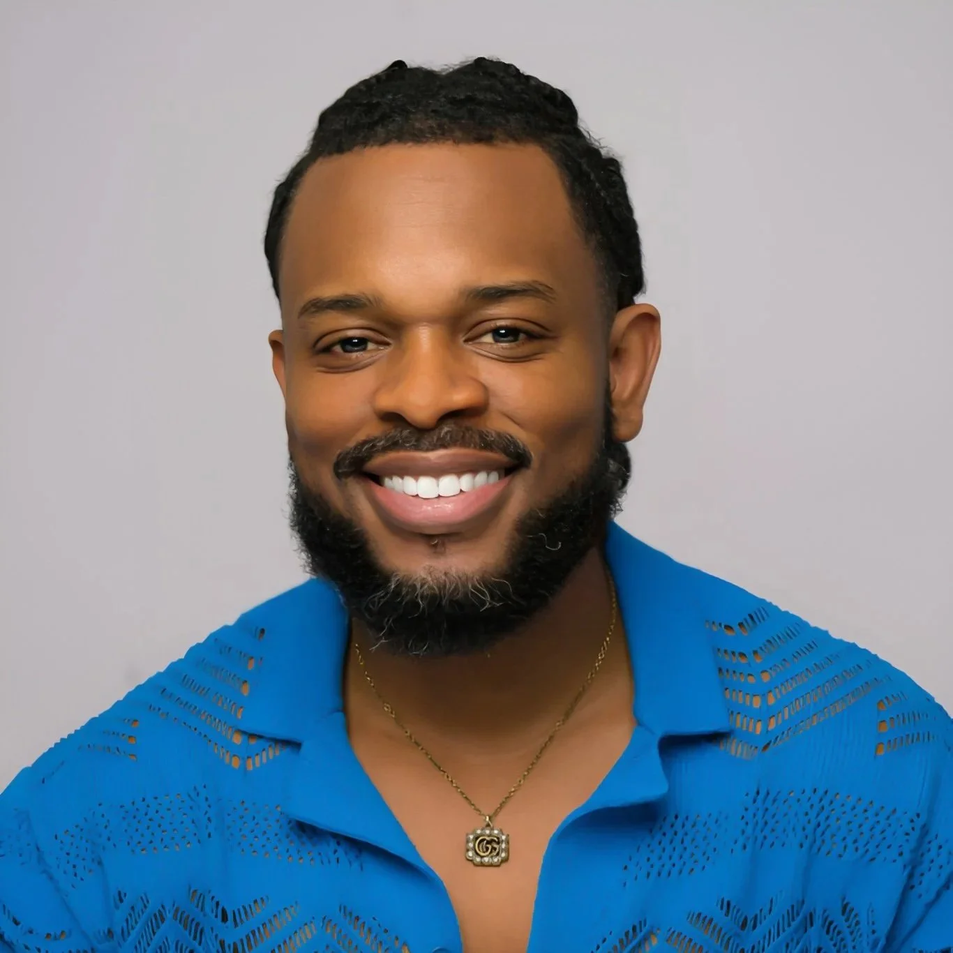 Portrait of a smiling man with a beard, wearing a blue shirt with cutout details, gold necklace, and a pendant.