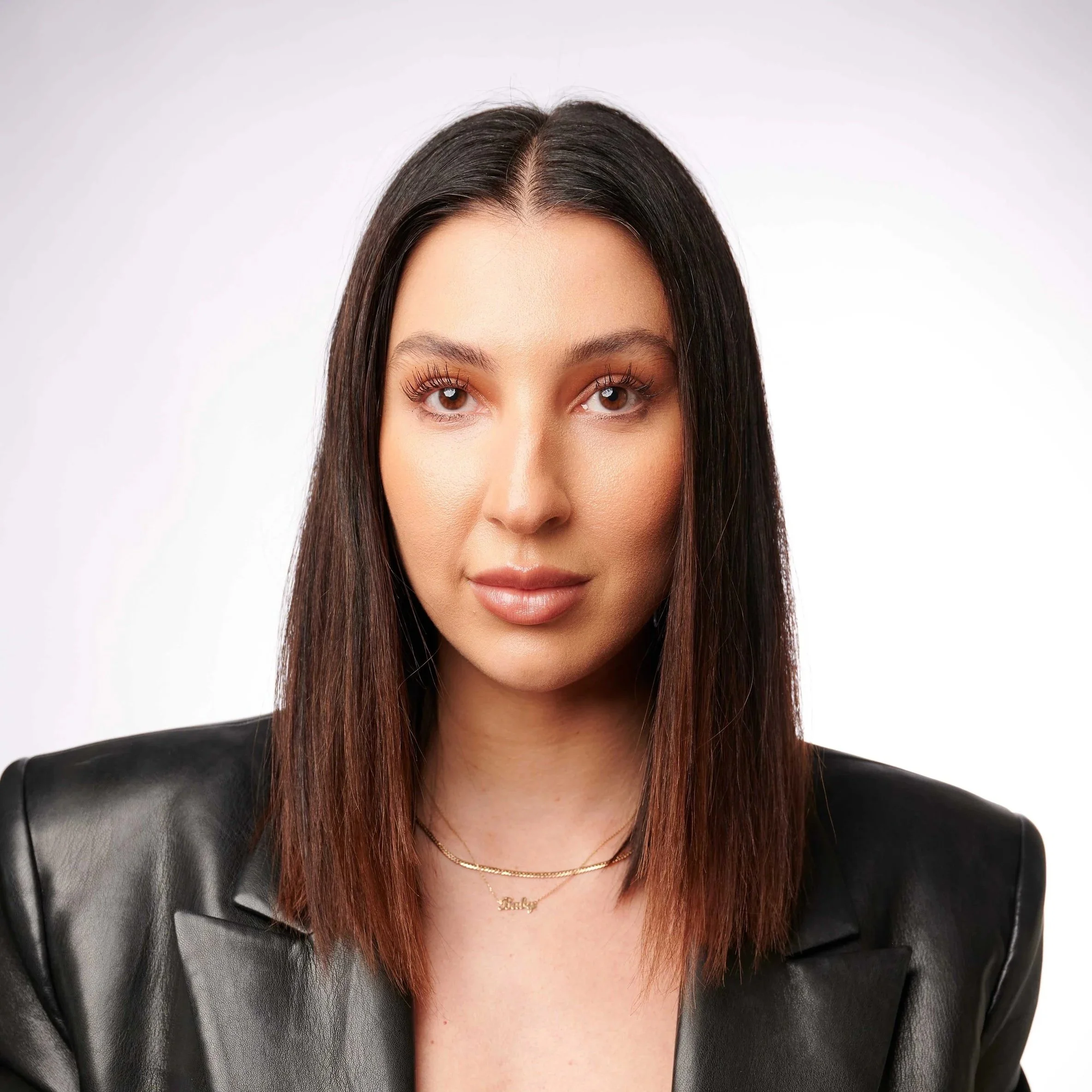 A young woman with shoulder-length brown hair, wearing a black leather jacket and a gold necklace, looking directly at the camera with a neutral expression.