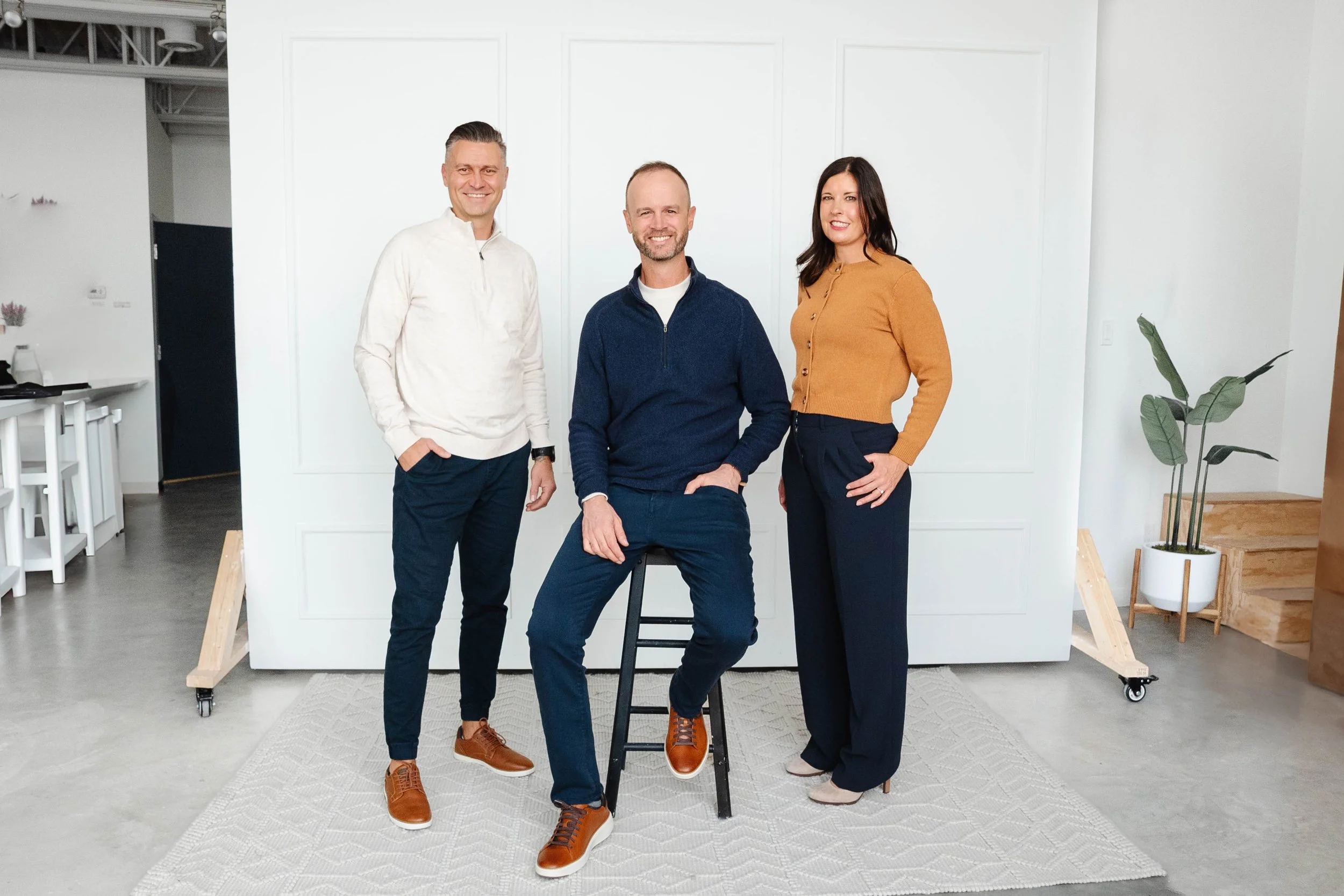 Three people posing indoors in front of a plain white wall. One man is sitting on a stool, flanked by a man and a woman standing on either side. The man sitting has a beard and is smiling, wearing a navy blue sweater and blue pants. The man standing to the left has short hair and is wearing a cream-colored sweater and dark pants. The woman standing to the right has long dark hair and is wearing a mustard-colored cardigan and dark pants. There is a plant on the right side in a white pot, and some furniture and decor visible in the background.
